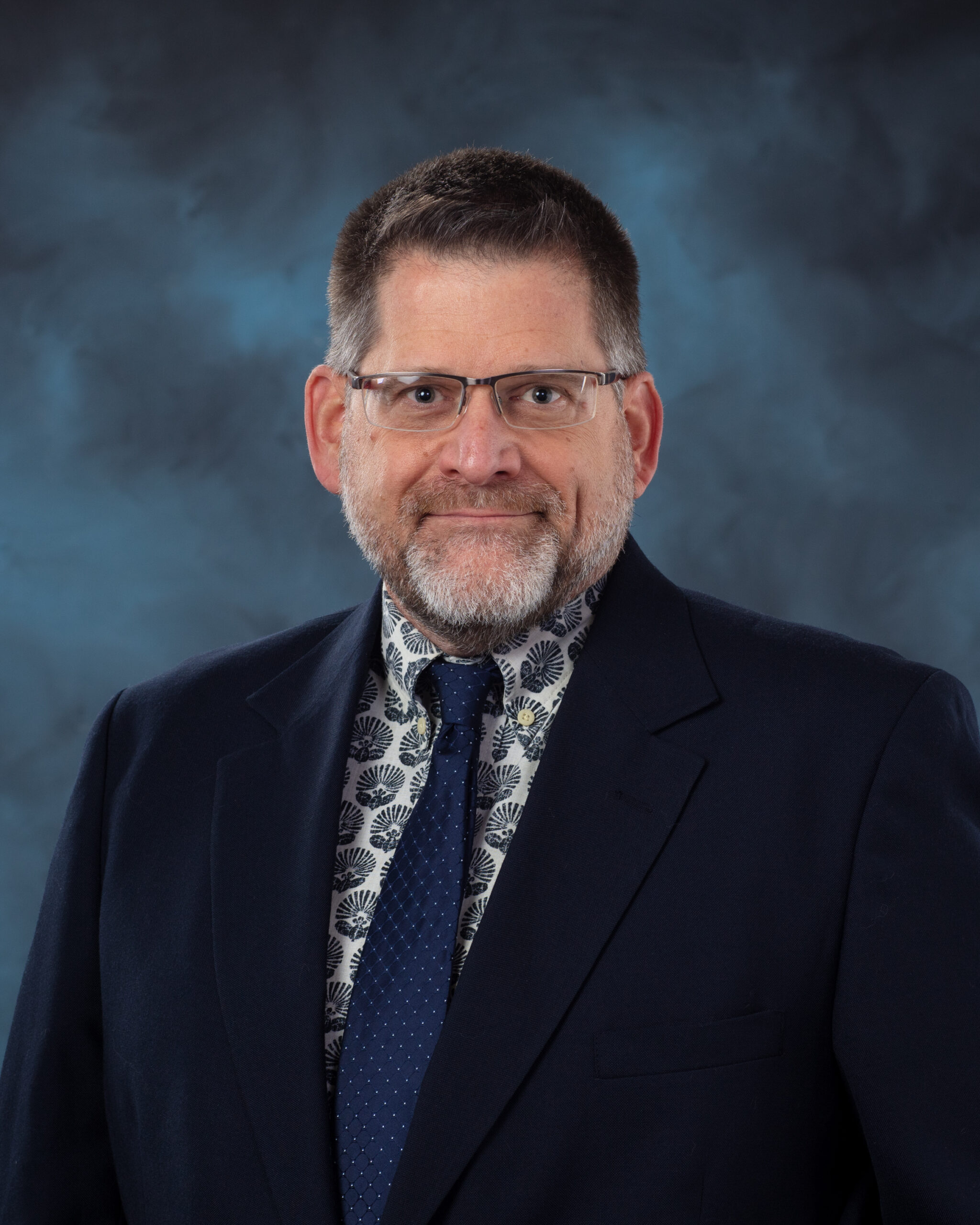 A man wearing glasses, a navy suit, and a patterned shirt with a dotted tie poses confidently with arms crossed against a dark blue textured background.