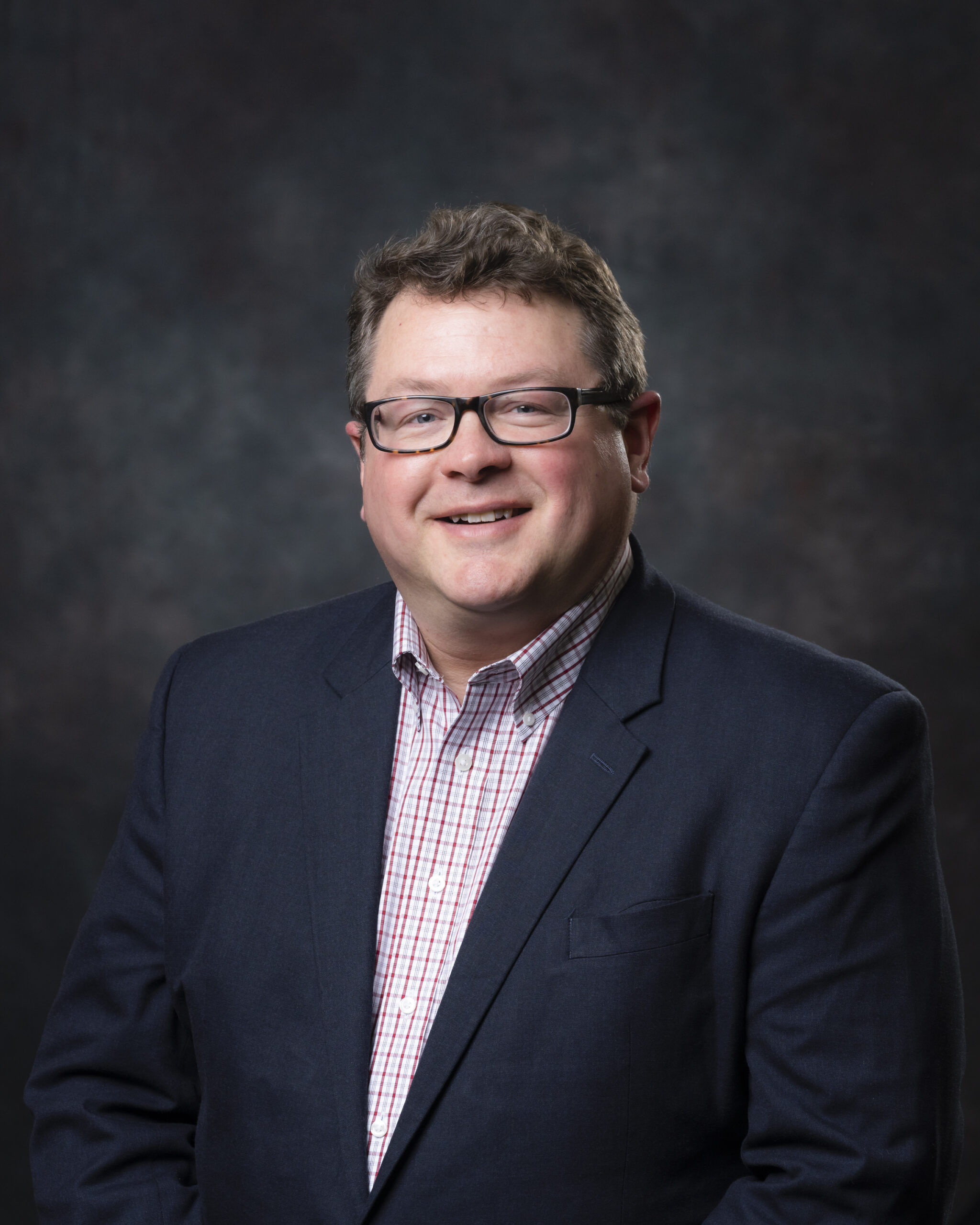 ORNL program manager Bill Peter is posing for a photo with a dark grey background and black suit with glasses