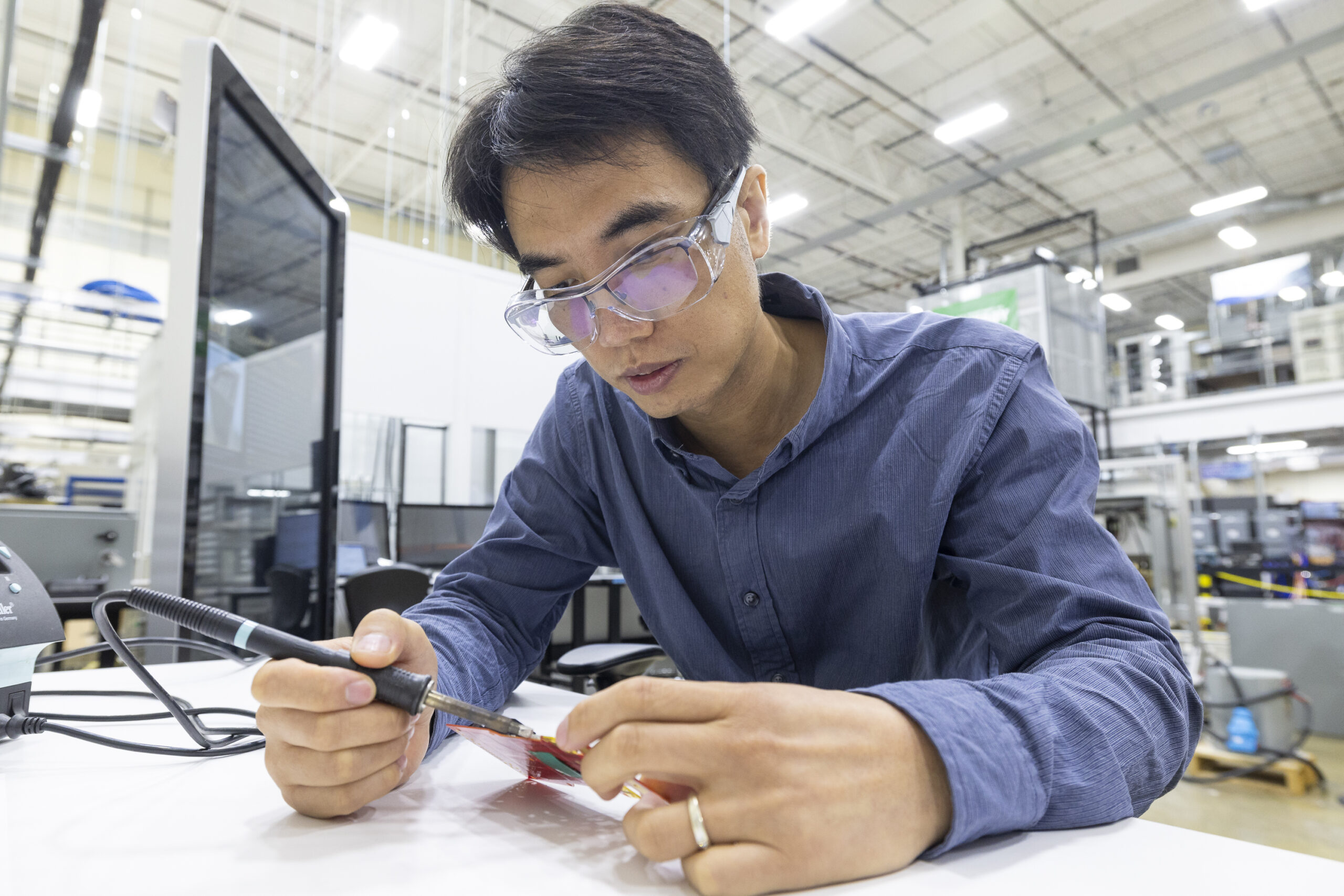 This photo is of a male scientist sitting at a desk working with materials, wearing protective glasses.