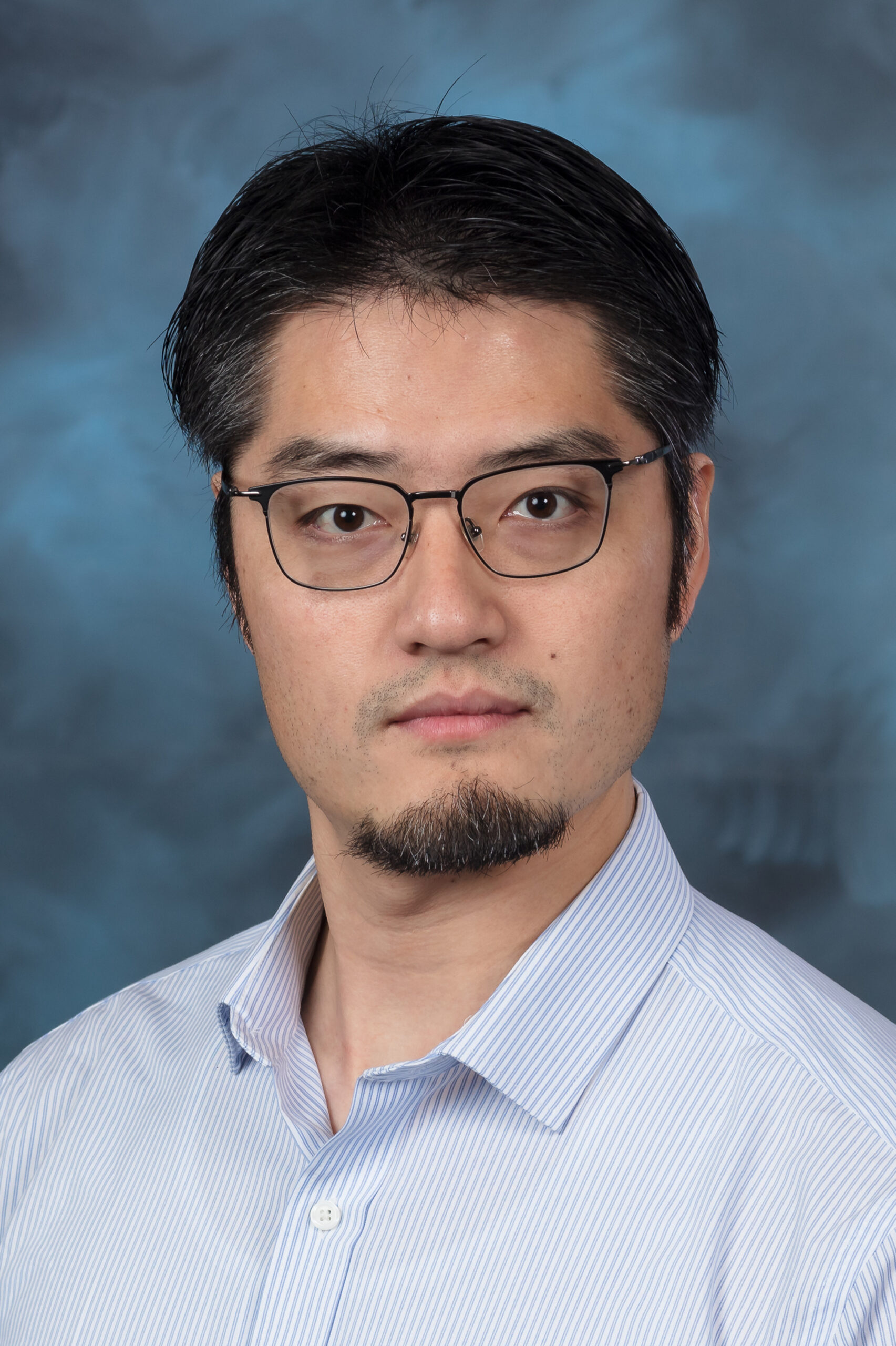 Man with dark brown/black hair with glasses is wearing a light blue button up, smiling for a photo with a blue backdrop.