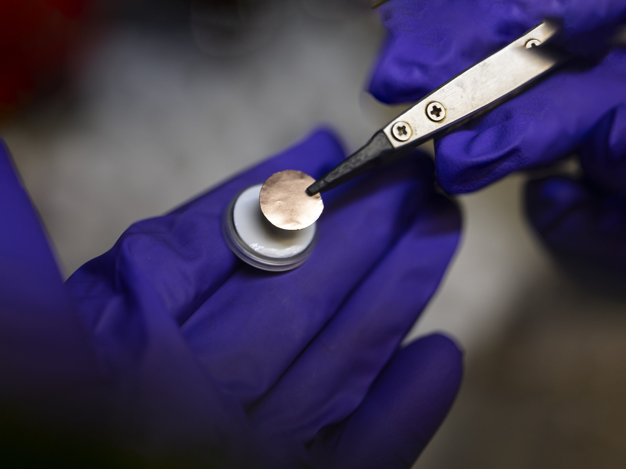 A researcher holds a test coin cell battery with purple gloves and a set of what looks like tweezers