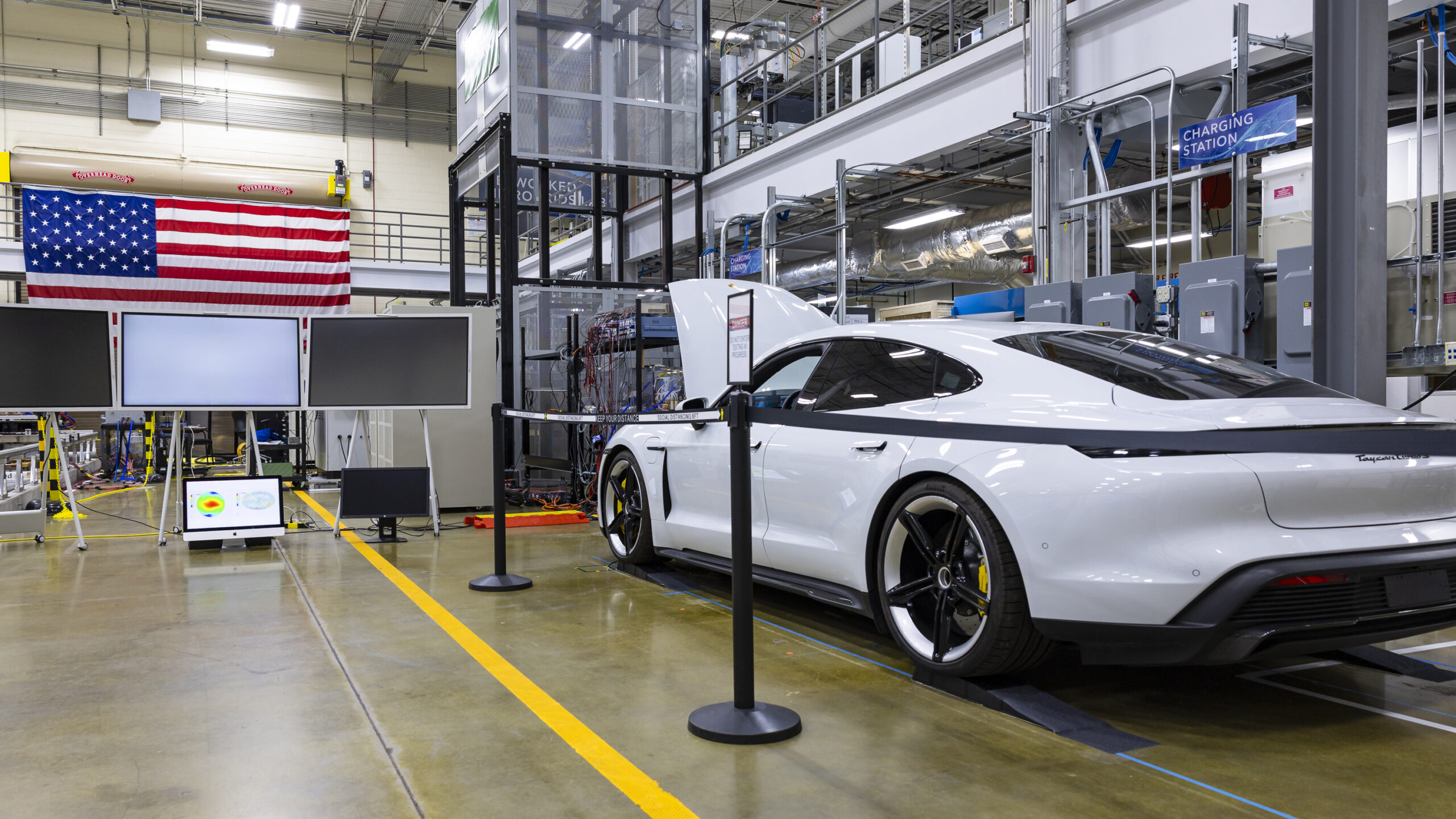 White car (Porsche Taycan) with the hood popped is inside the building with an american flag on the wall.
