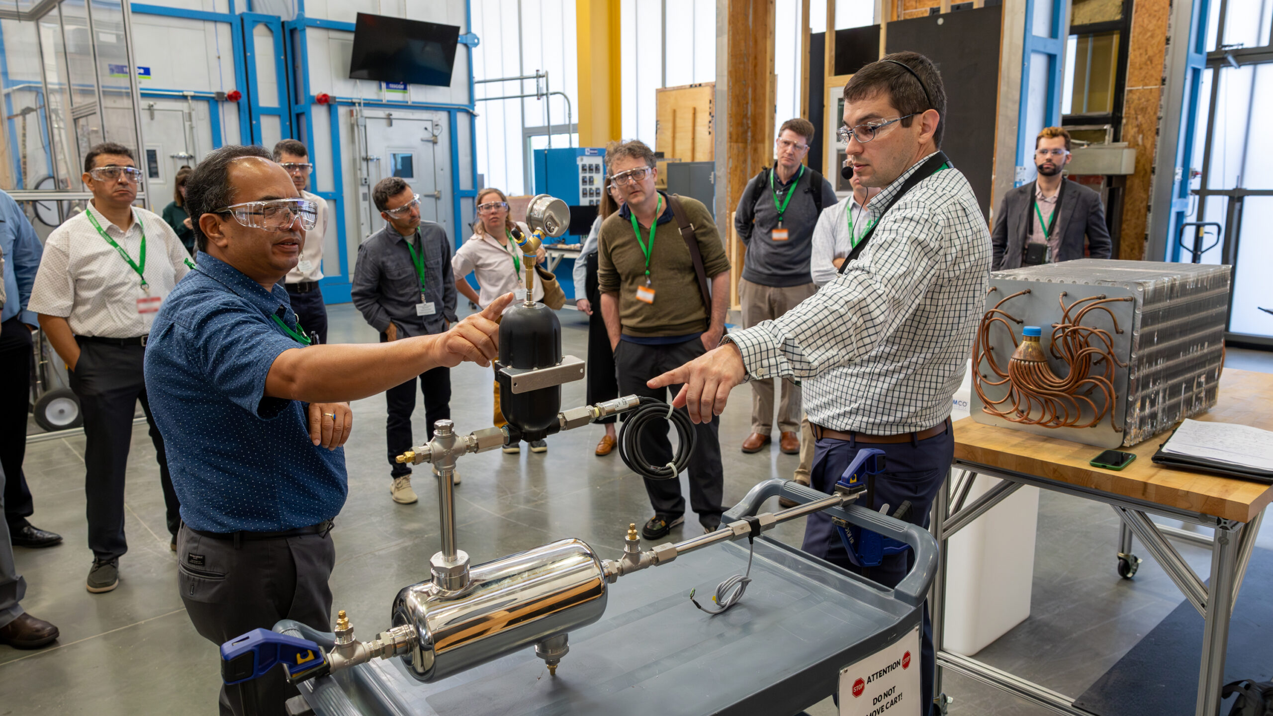 Two scientists are standing in the lab pointing at an object on the table, both wearing glasses.
