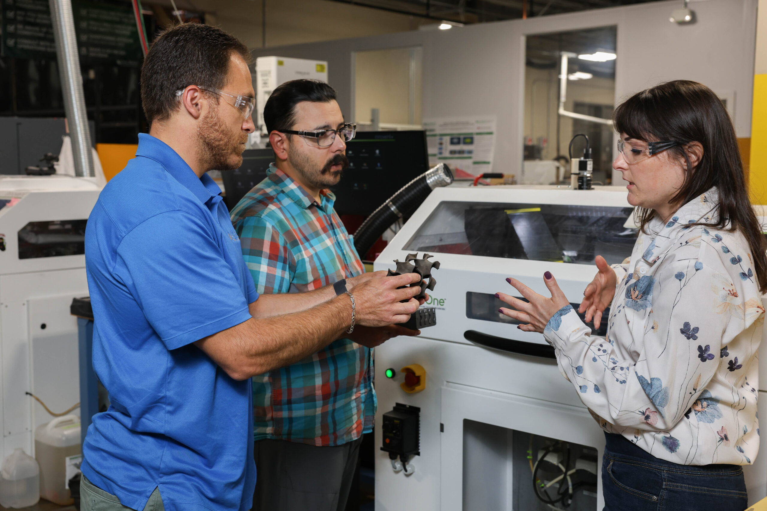 From left, Corson Cramer, Trevor Aguirre and Amy Elliott discuss the silicon carbide gyroid component, which was 3D printed using the binder jet printer displayed in the background.