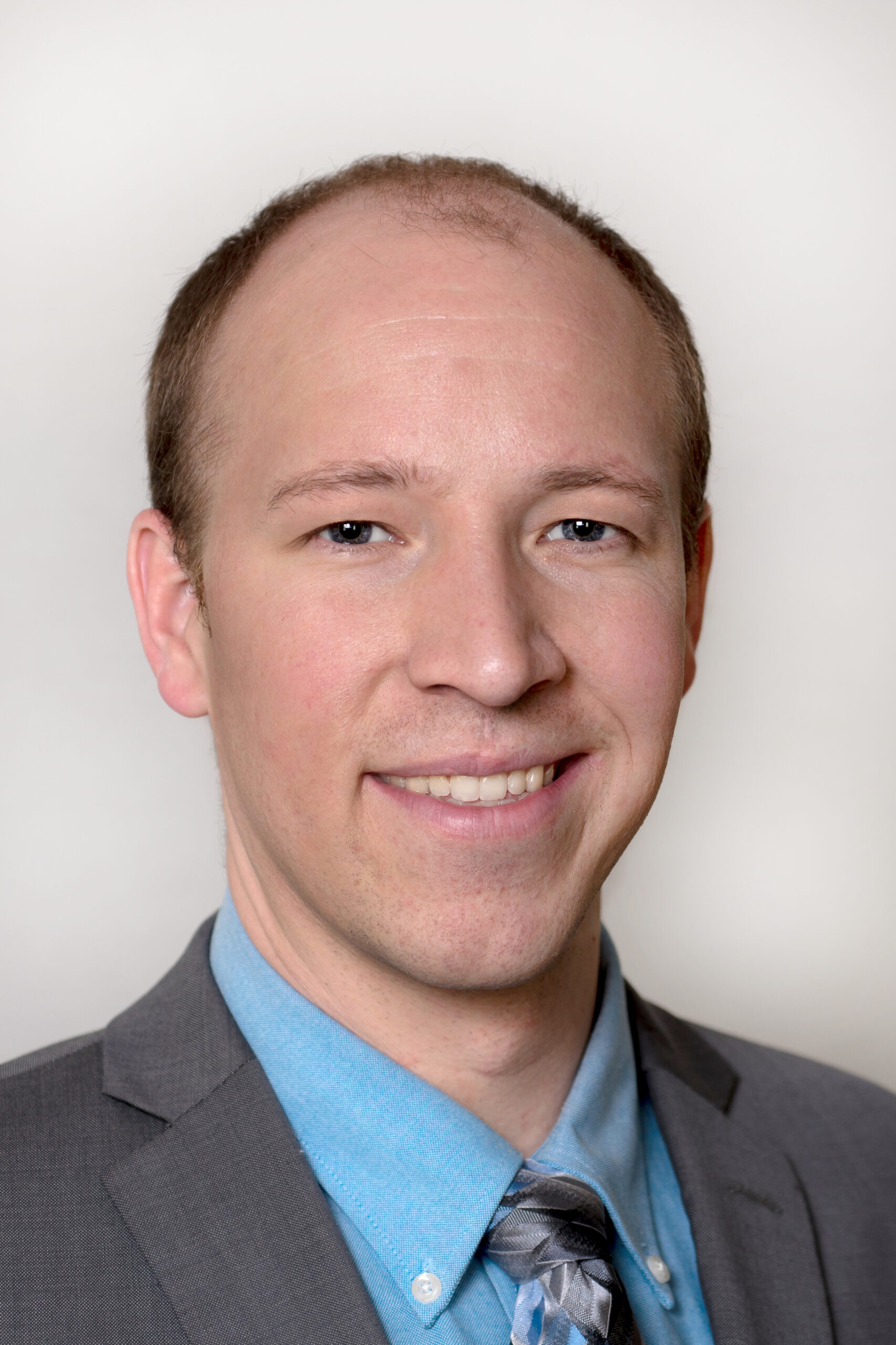 Man in a grey suit and blue button up with a grey tie smiles for a photo with a black backdrop. 