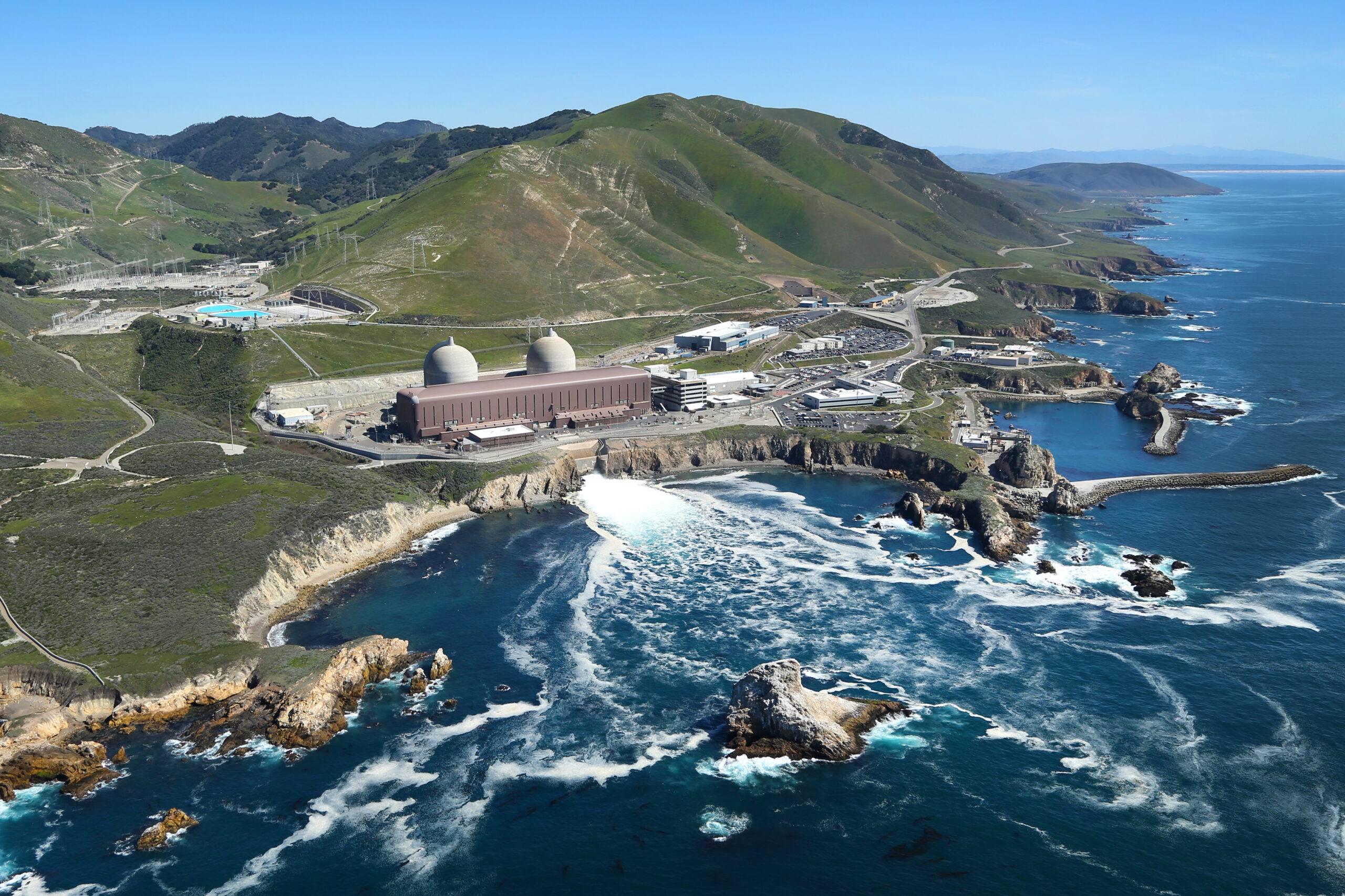 An aerial view of the Diablo Canyon Nuclear Power Plant situated on a rugged coastline, with two domed reactor buildings overlooking cliffs, waves, and the Pacific Ocean, surrounded by green hills and facility structures.