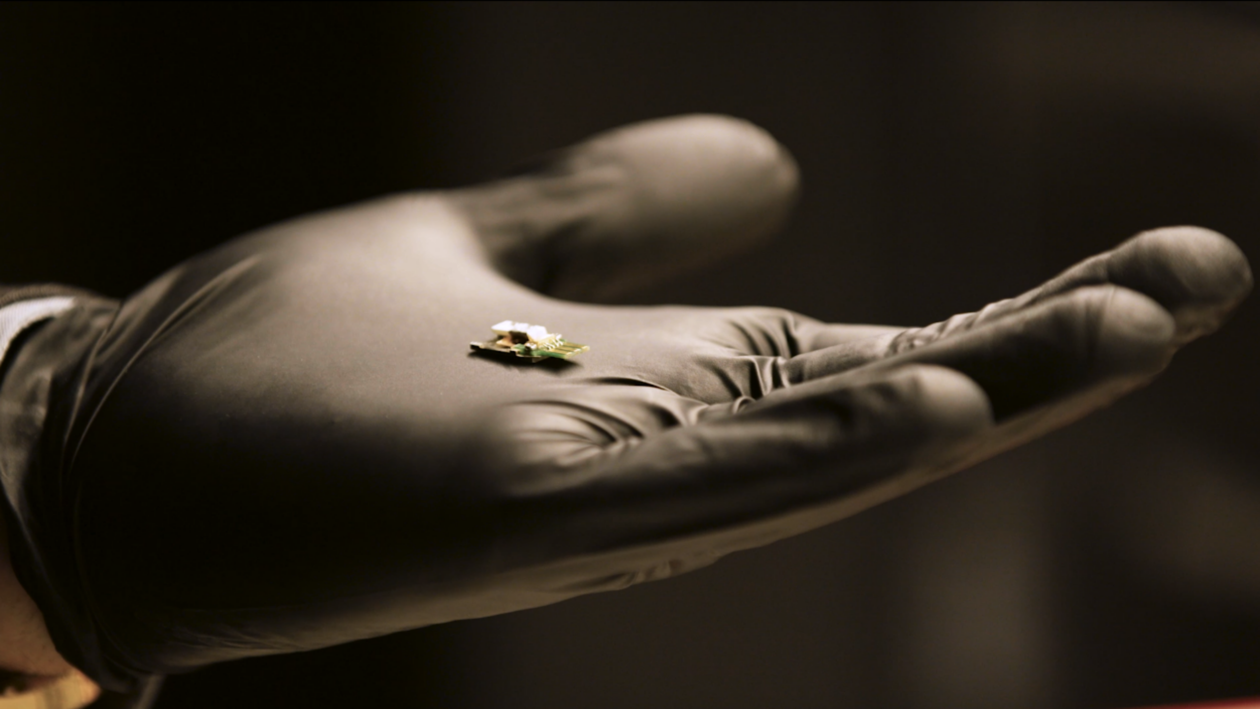 A hand in a black glove holds a heater platform designed to deliver atomized material to a sample