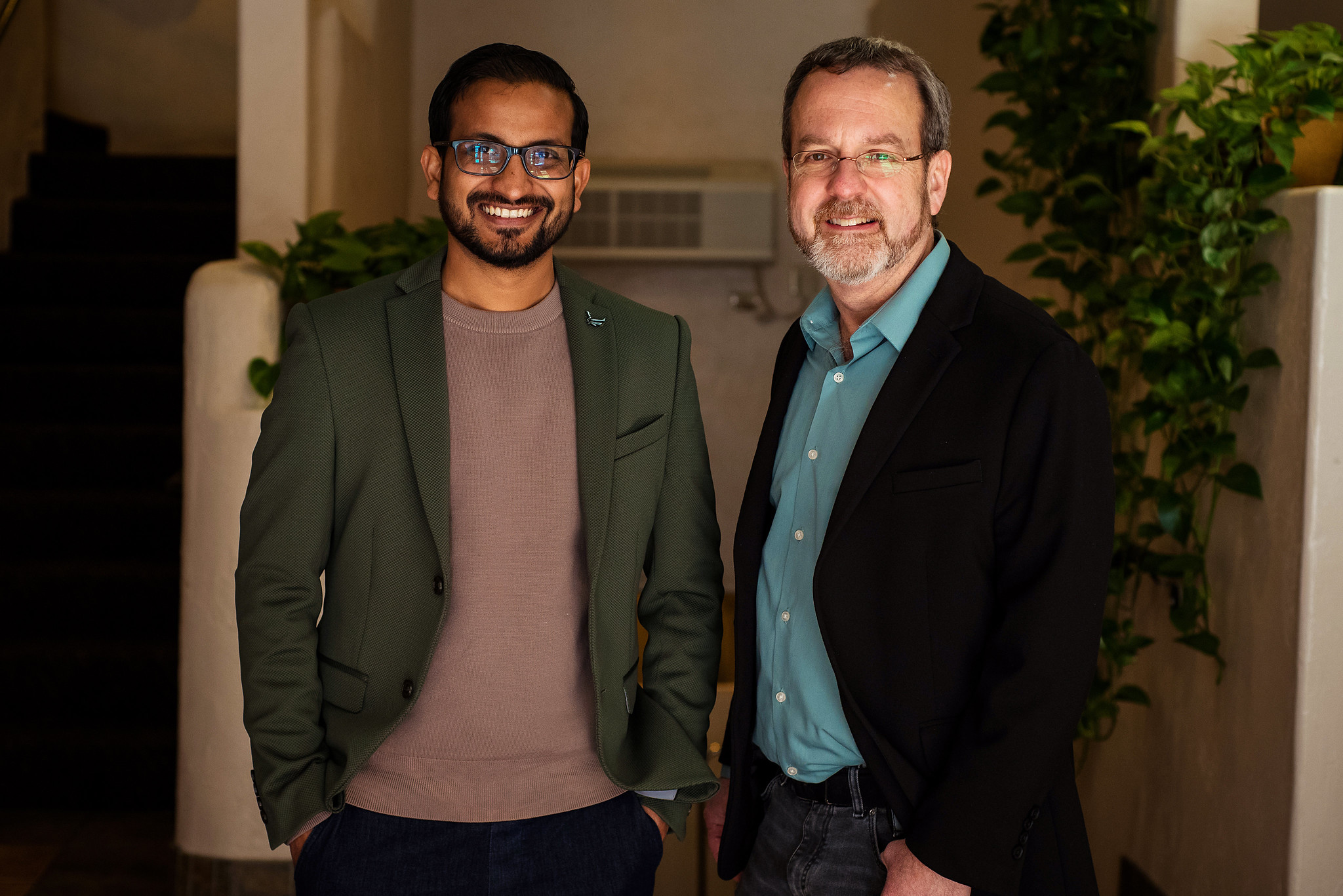 Two men standing in a hallway with a plant behind them. The man on the left is wearing a green jacket and glasses whereas the man on the right is wearing a teal button down and a black suit jacket.