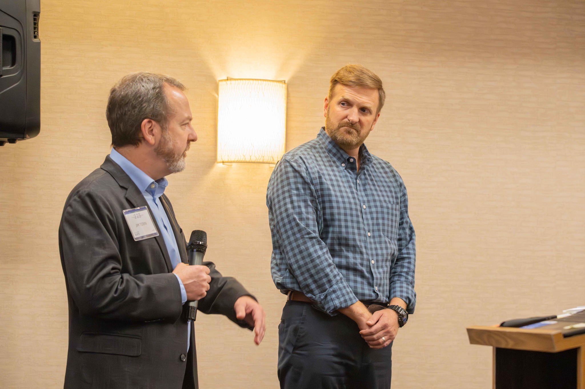 Two men are pictured here. Man on the left is talking with a microphone in a grey suit with blue shirt.