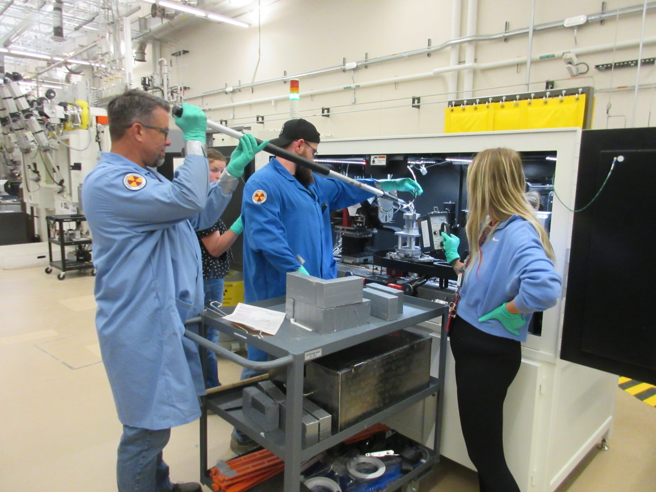 A picture containing indoor, person, working, preparing - sticking a rod into a chamber, wearing blue lab coats
