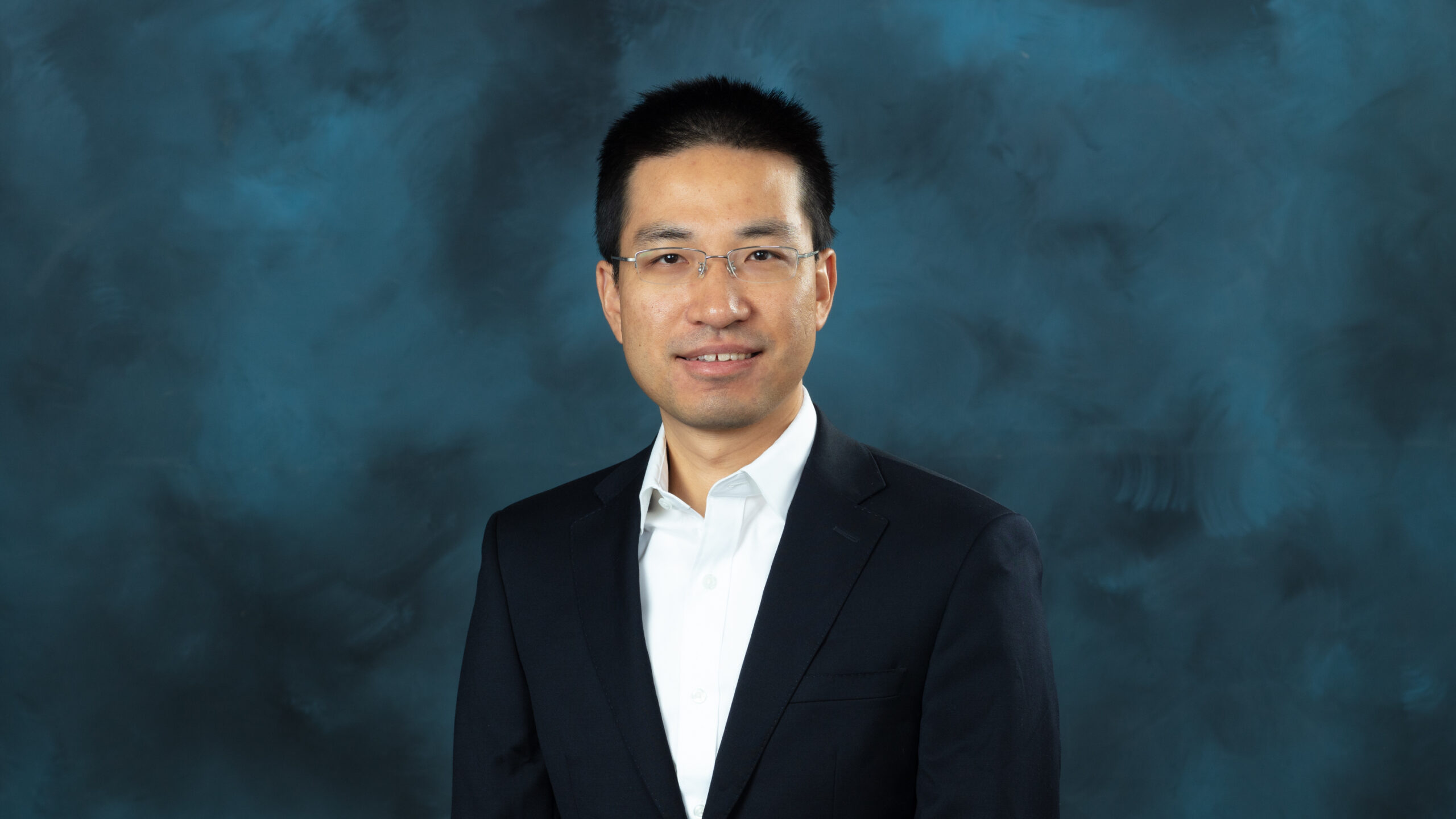Professional headshot of a man with short black hair and glasses wearing a white shirt and dark suit jacket, against a blue-gray background.