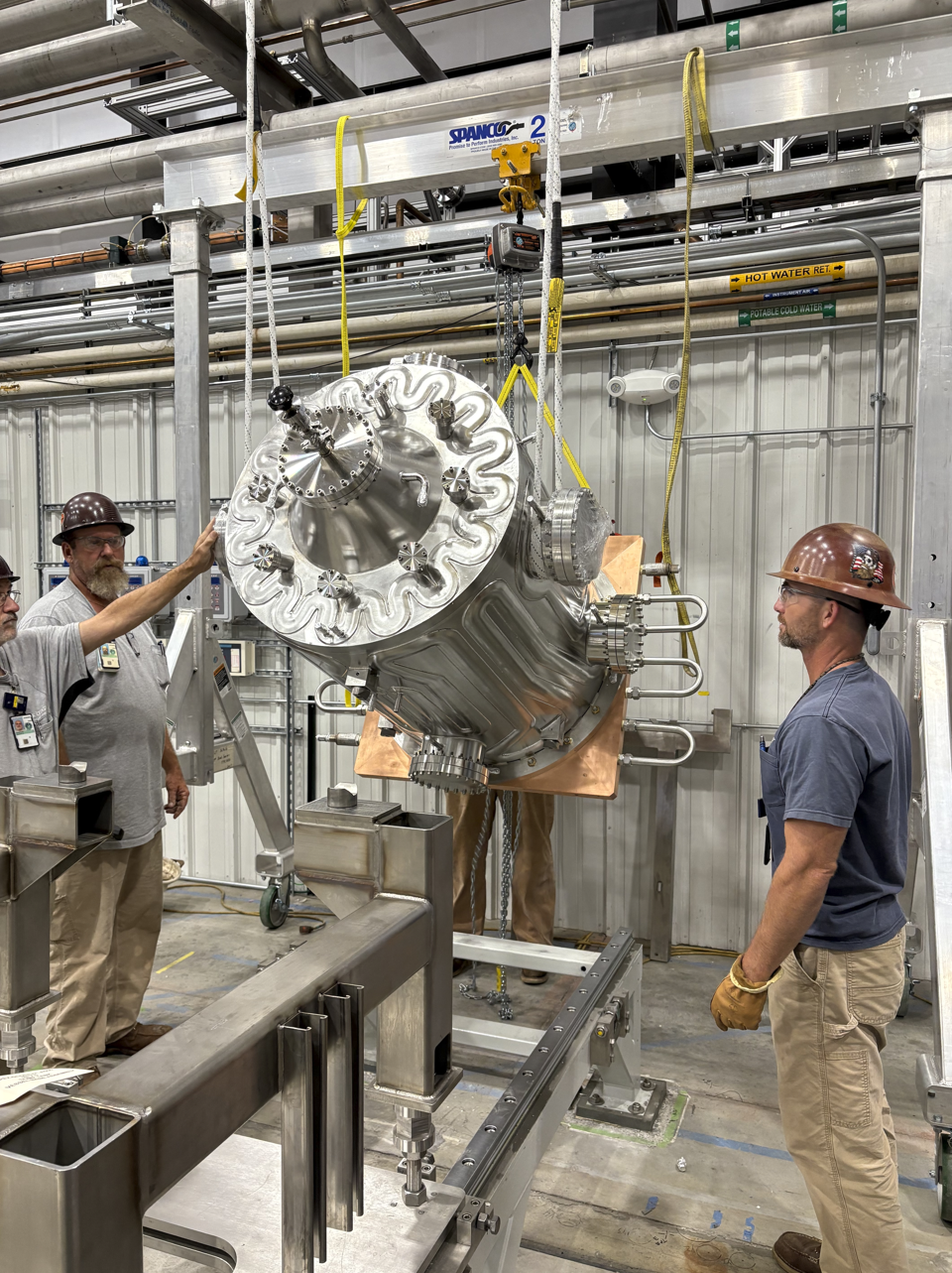 Several technicians wearing hard hats and gloves guide a large, suspended stainless-steel vacuum vessel into position using lifting straps and a hoist system. The vessel has a copper mounting plate and is being aligned with a metal support structure inside an industrial facility.