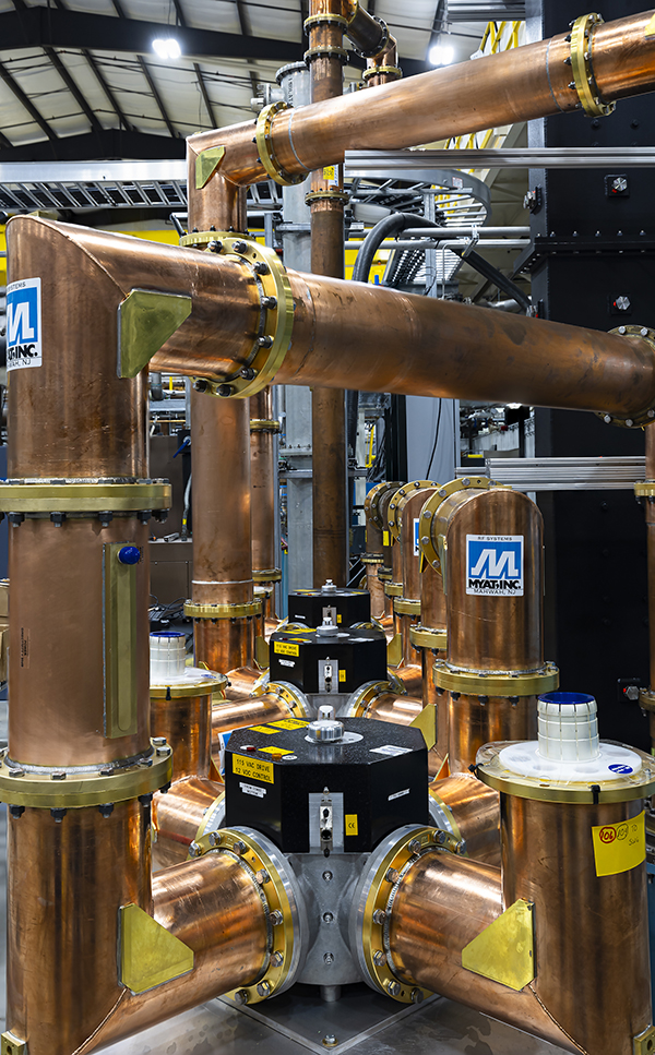 A close-up of a copper RF waveguide network with multiple interconnected rectangular copper pipes and vacuum components. Several black RF power combiners or switches are mounted at junctions, with manufacturer labels and safety markings visible.