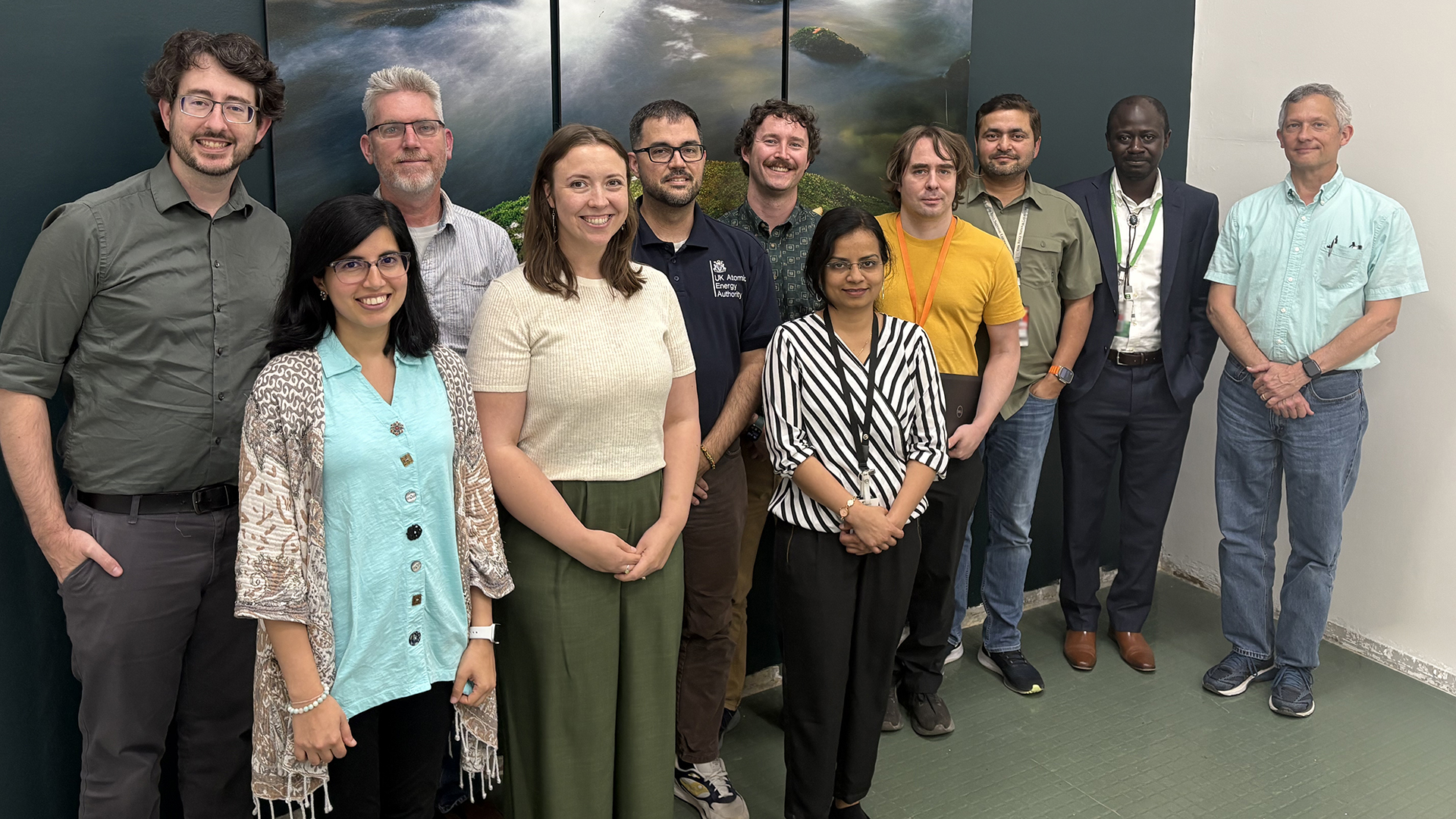 A group of eleven people stand indoors in front of a large nature-themed wall mural. The group includes men and women of diverse ages, smiling and posing for a team photo.