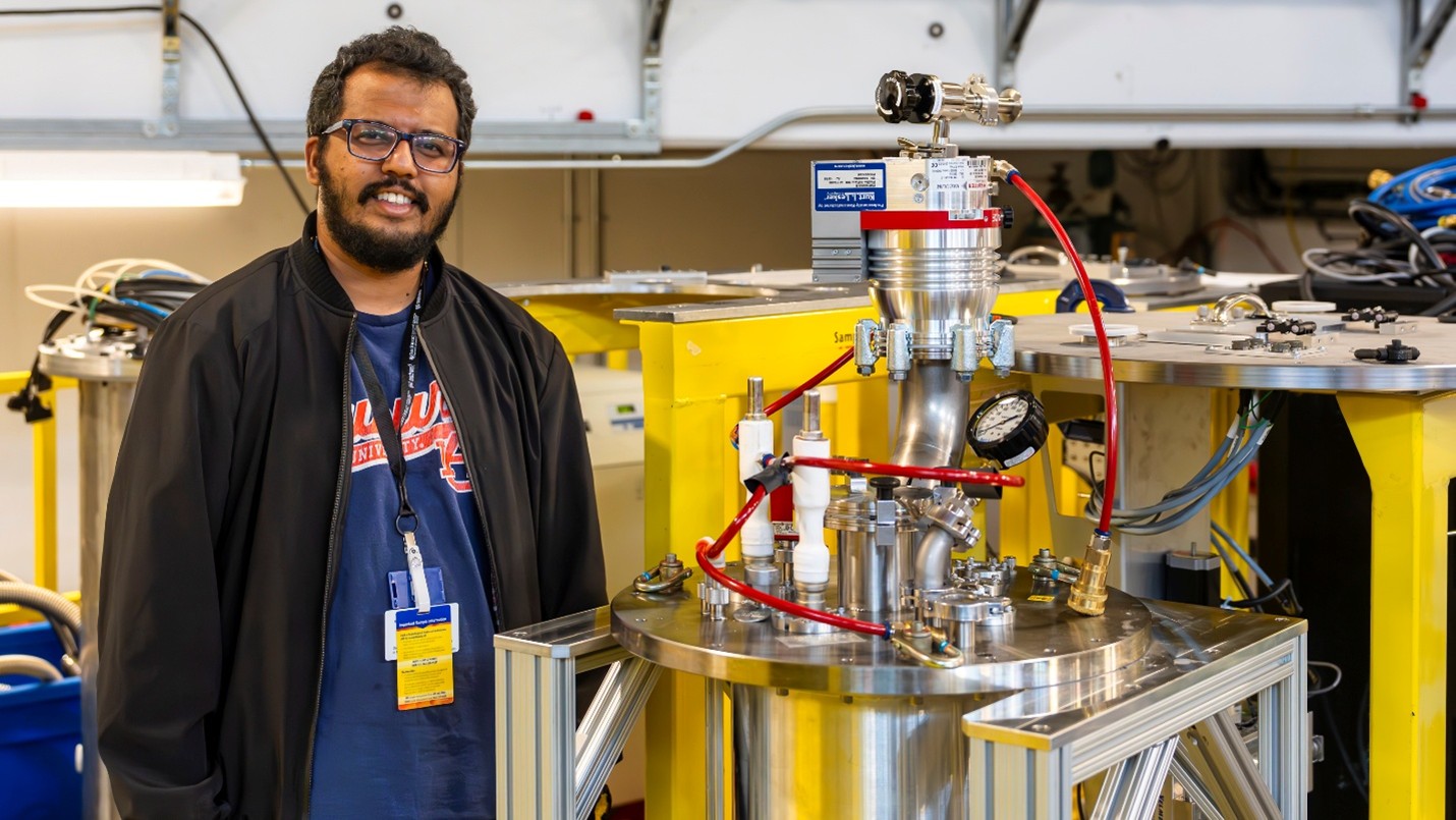 A researcher stands beside scientific equipment with yellow framework and metal tubing in a laboratory at Oak Ridge National Laboratory.