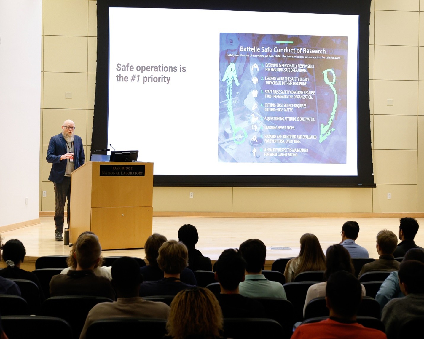 A speaker stands at a podium labeled “Oak Ridge National Laboratory,” presenting a slide about safe operations to an audience seated in an auditorium.