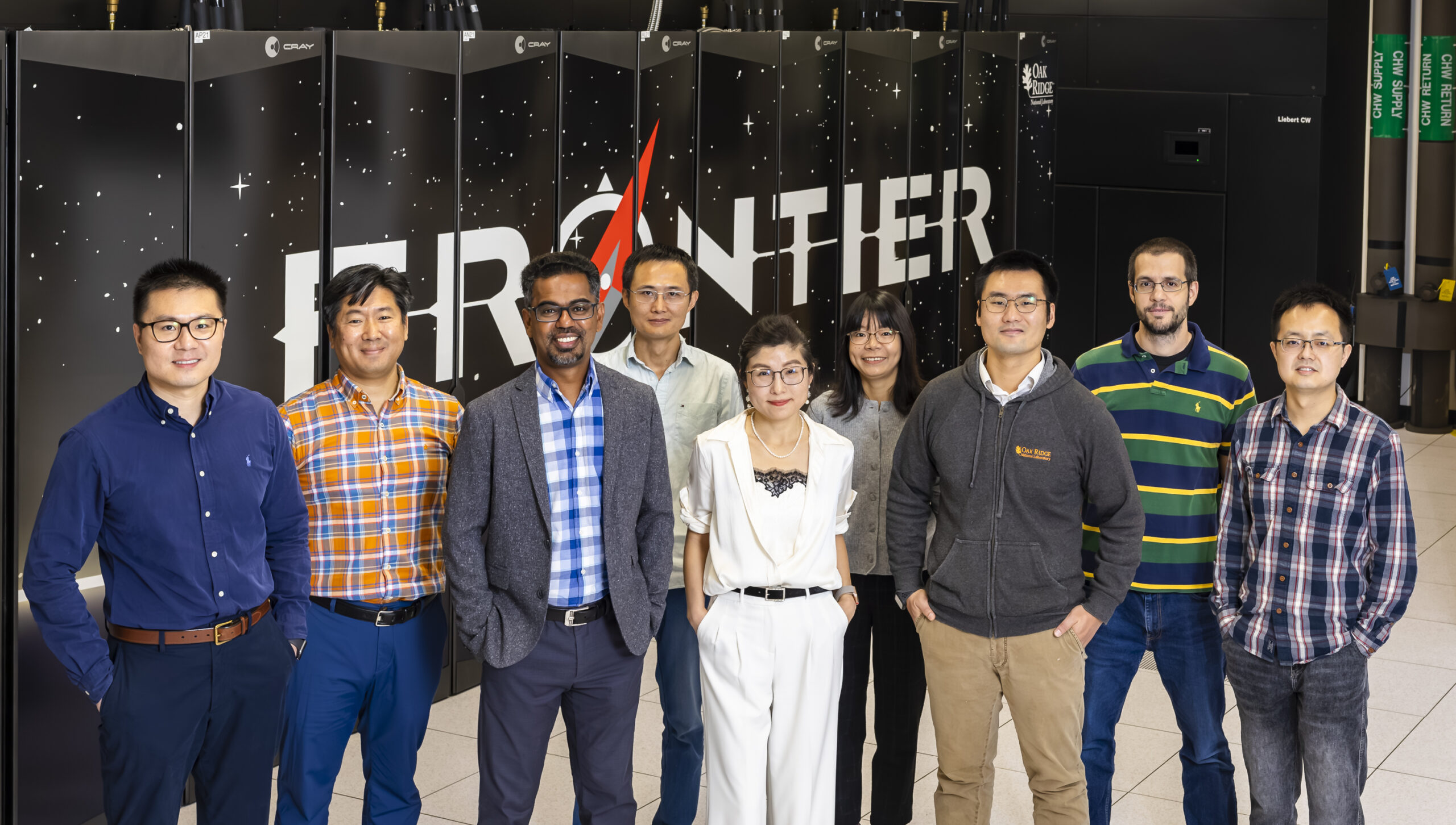 Pictured here are 9 scientists standing in a line in front of the frontier supercomputer logo/computer