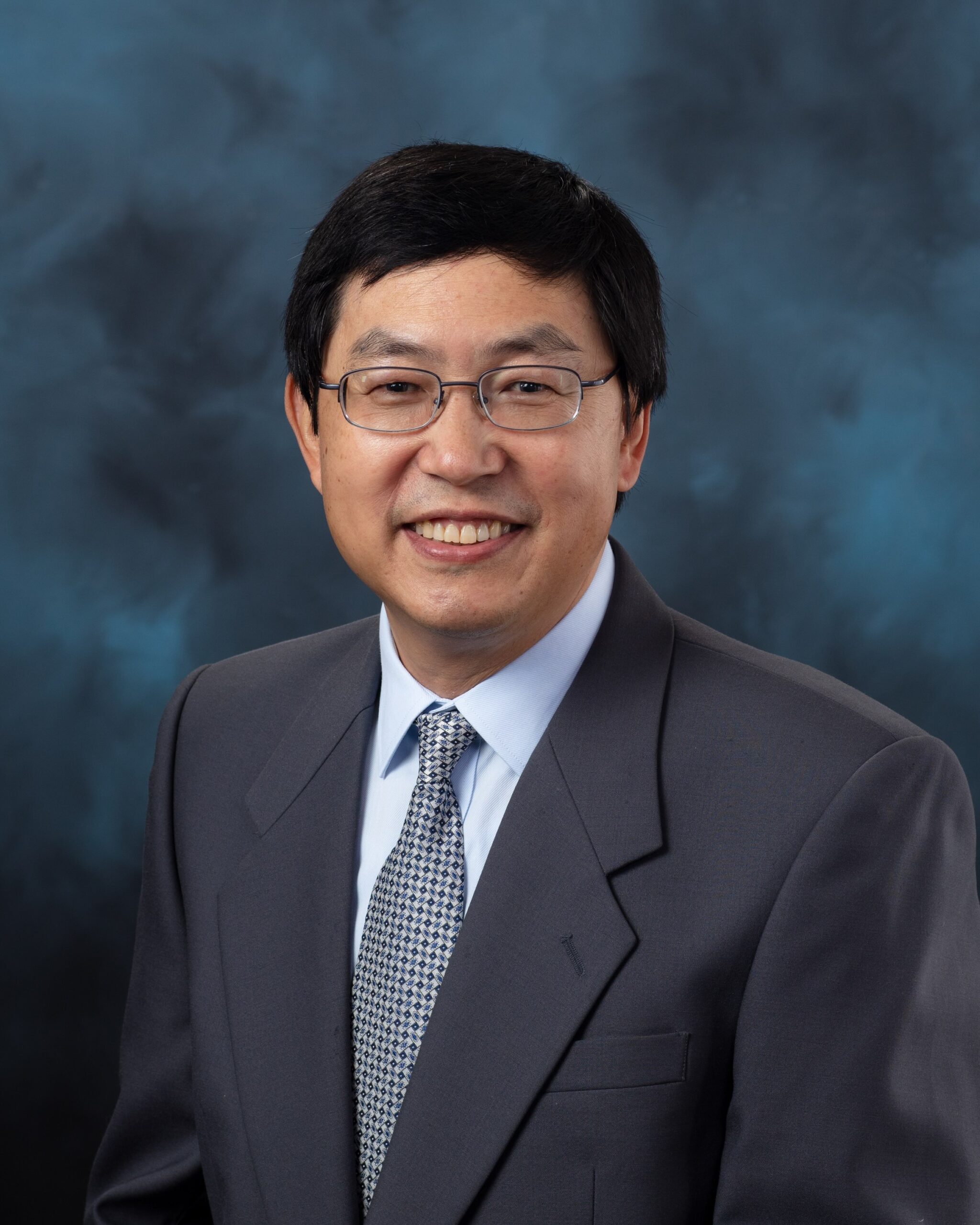 Professional portrait of a man in a dark suit and patterned tie, smiling slightly against a dark blue studio background.