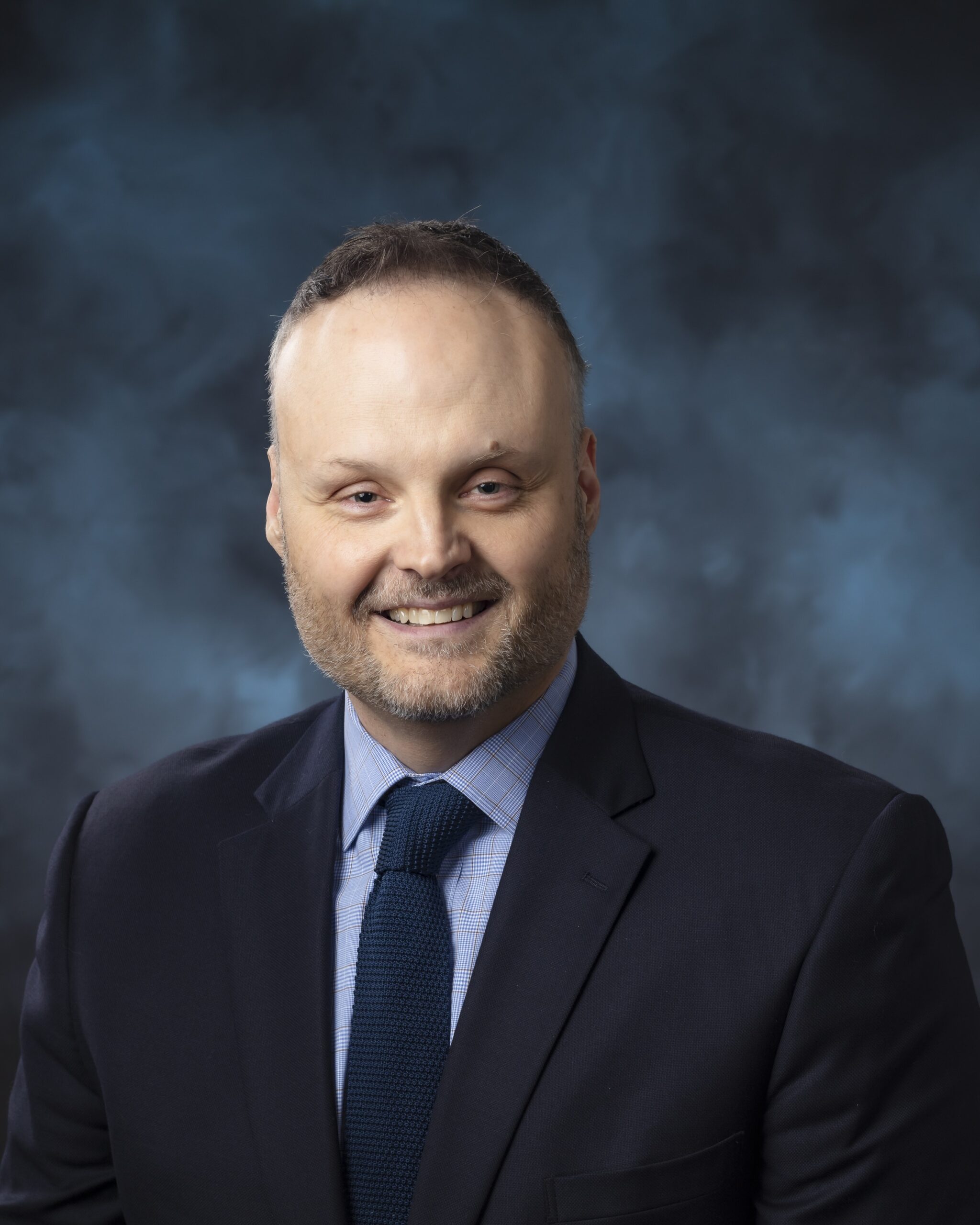 Professional portrait of a man with light hair and trimmed beard, wearing a navy suit, light blue checkered shirt, and dark blue tie, smiling against a blue-gray studio background.
