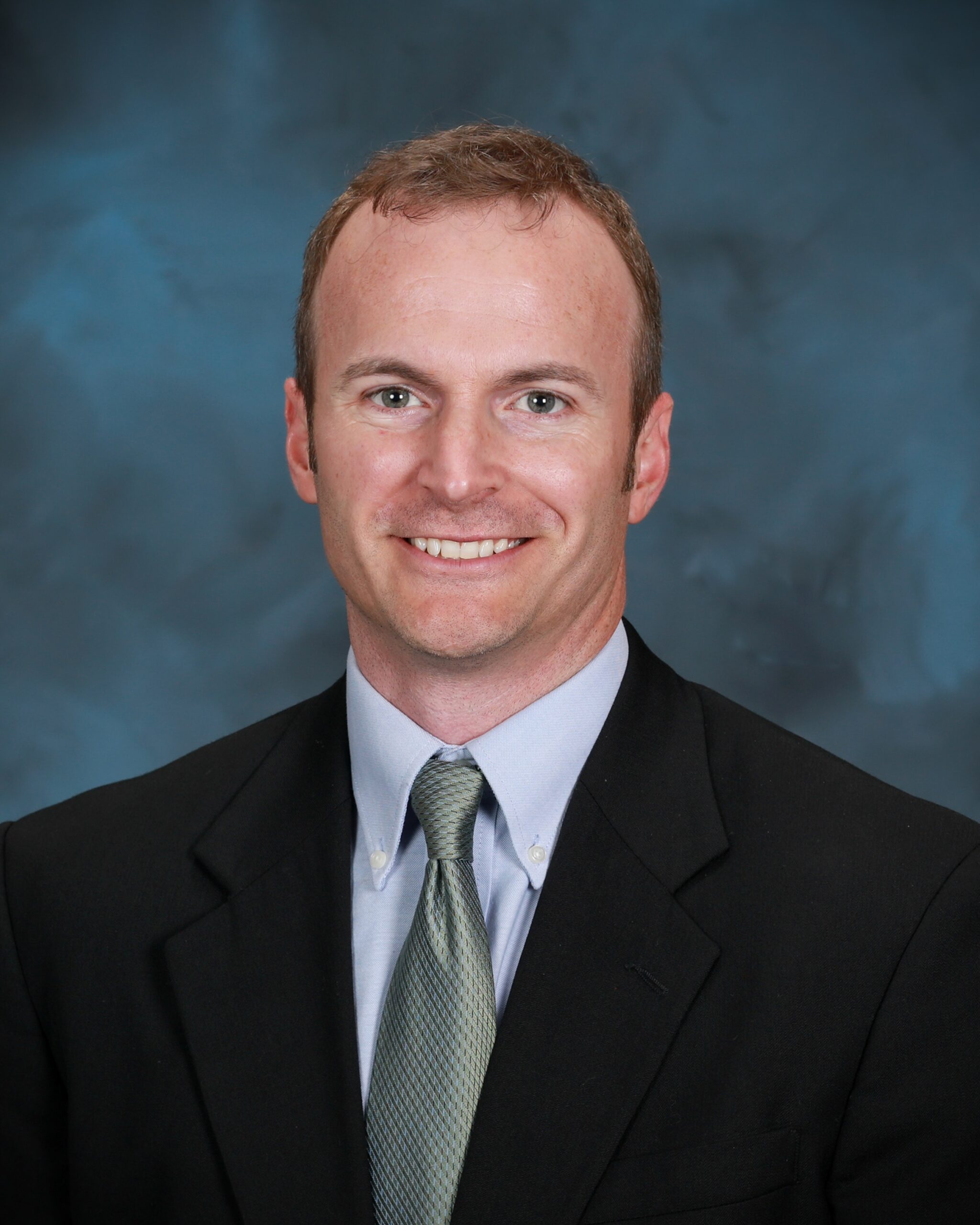 Professional portrait of a man wearing a dark suit, light blue shirt, and green tie, smiling against a dark blue studio background.