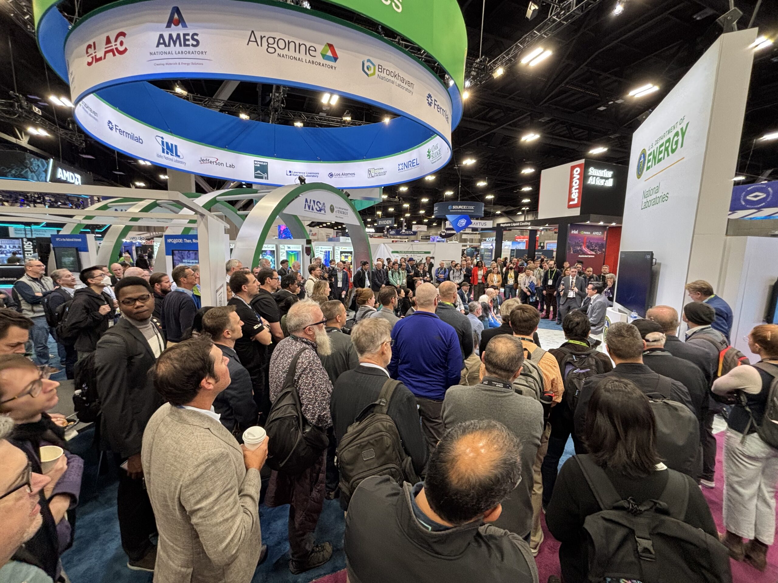 A large crowd of conference attendees gathers around a booth at a technology or scientific expo. Overhead signage displays logos of U.S. Department of Energy national laboratories such as SLAC, Ames, Argonne, Brookhaven, INL, and others. People stand closely together, many wearing badges and backpacks, while a presenter at the front speaks near a screen. The exhibition hall is filled with colorful booths, lighting rigs, and branded displays from various organizations.