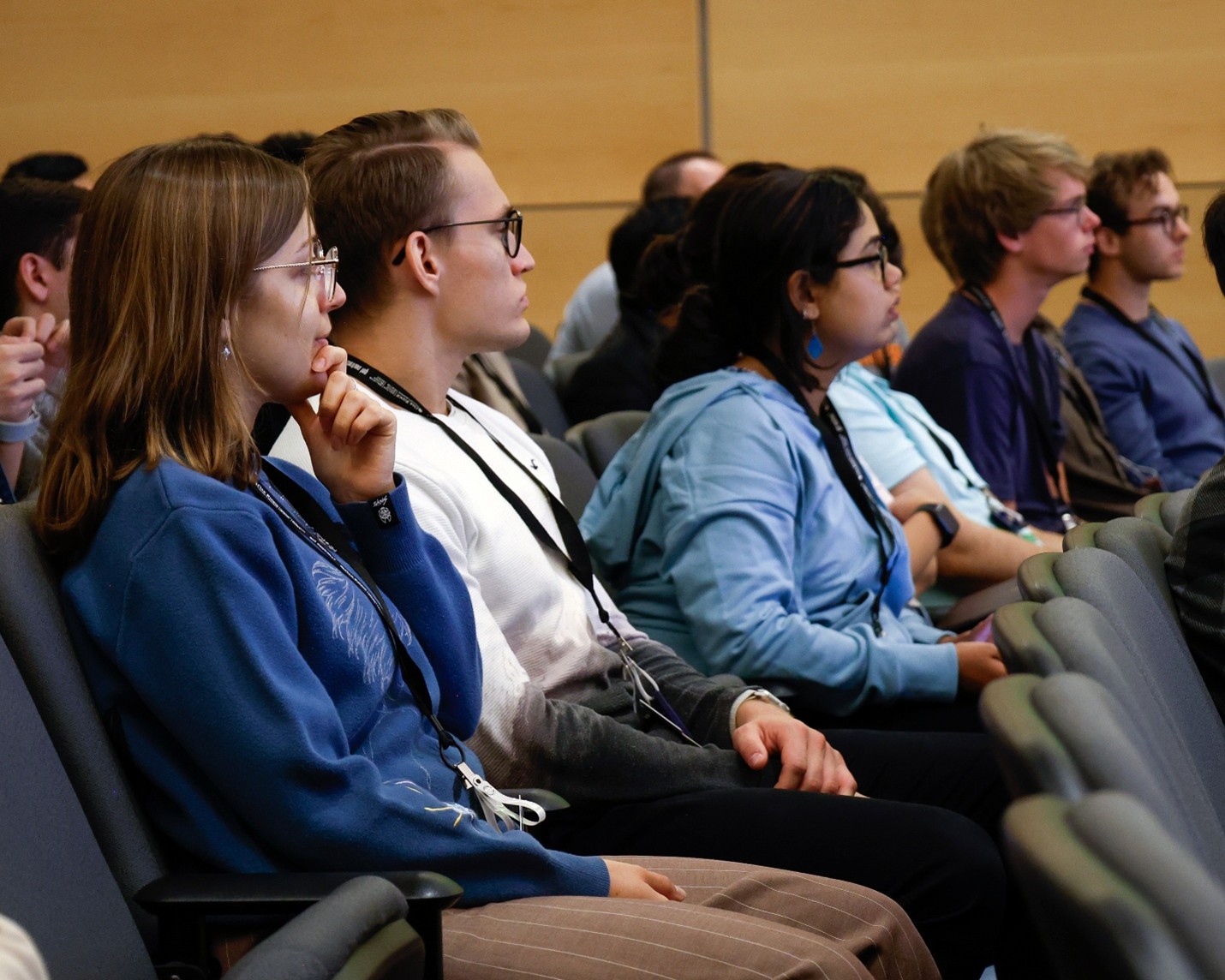 Students sit in an auditorium, attentively watching a presentation, some taking notes, wearing conference badges and casual attire.