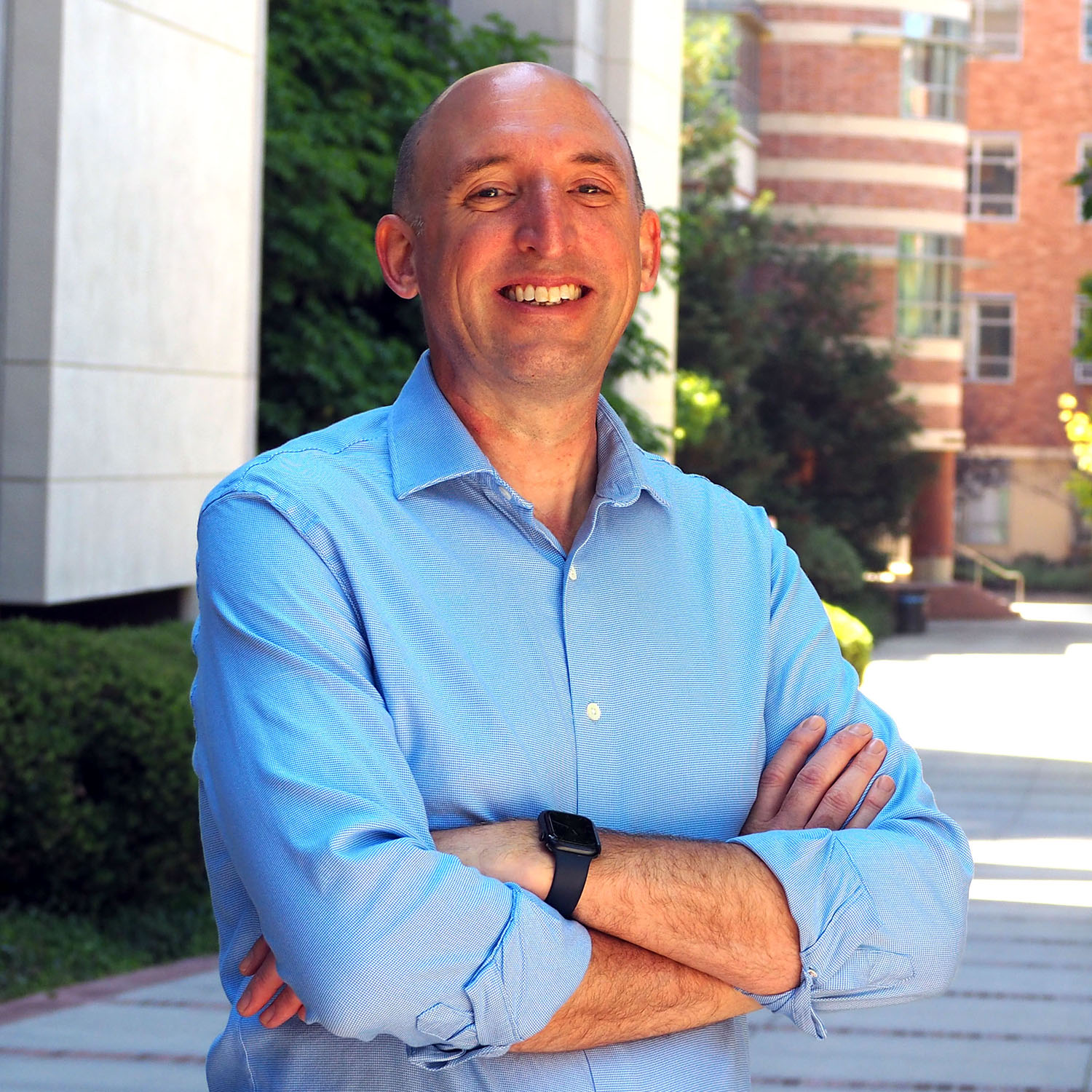 Man in blue button down shirt poses outside for a picture with his arms crossed.