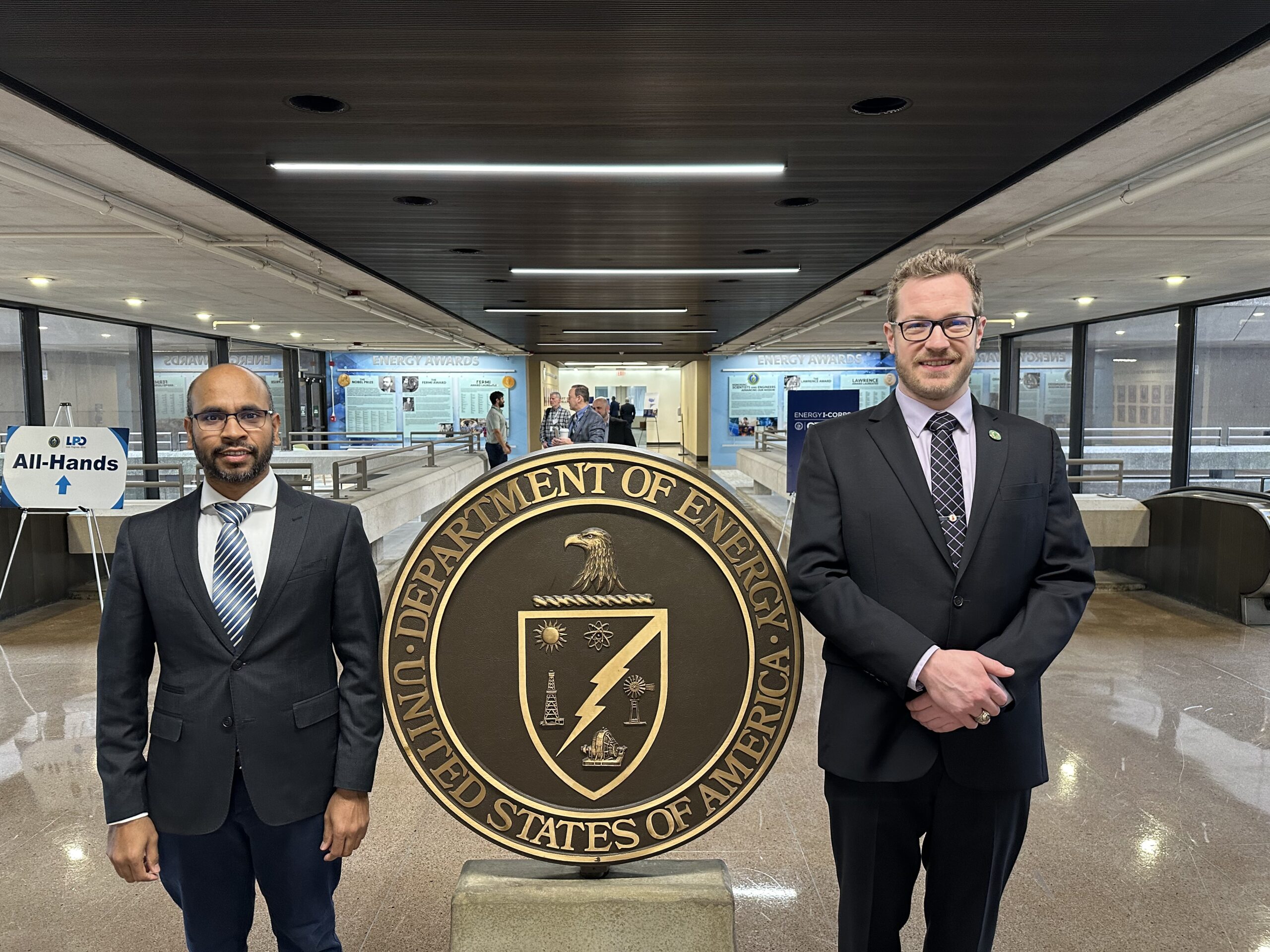 Two ORNL scientists are standing on each side of a large round logo.