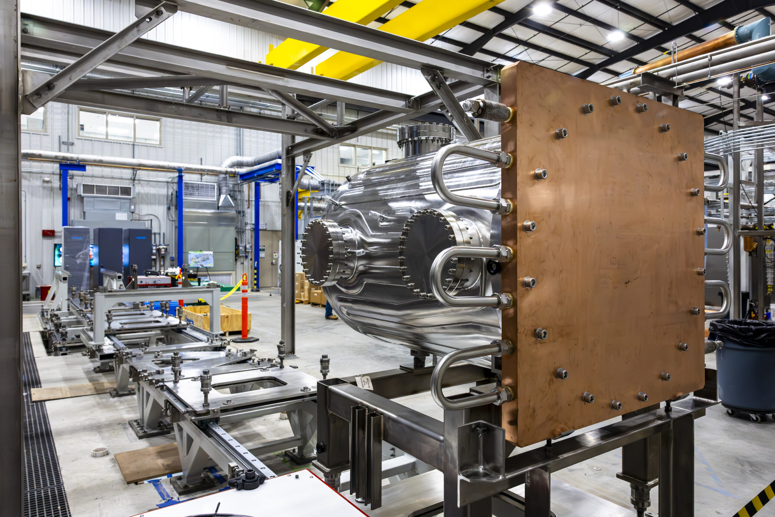 A large, polished stainless-steel vacuum chamber with flanged ports is mounted horizontally on a metal support frame in a high-bay laboratory. The chamber has a copper endplate with multiple bolts and is surrounded by industrial equipment, piping, and overhead cranes.