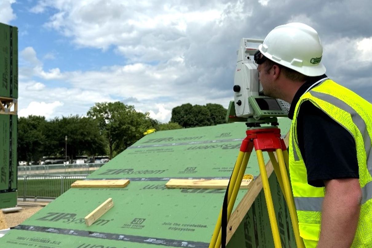 Male in hard hat and safety vest looks through tool for building.