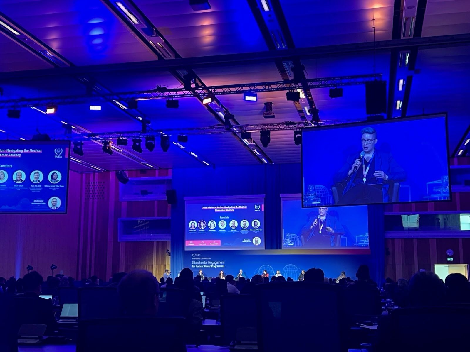 Gale Hauck speaks on a panel during the first annual International Atomic Energy Agency Stakeholder Engagement Conference with a blue hue and the background screen having 7 images of reasearchers in circles