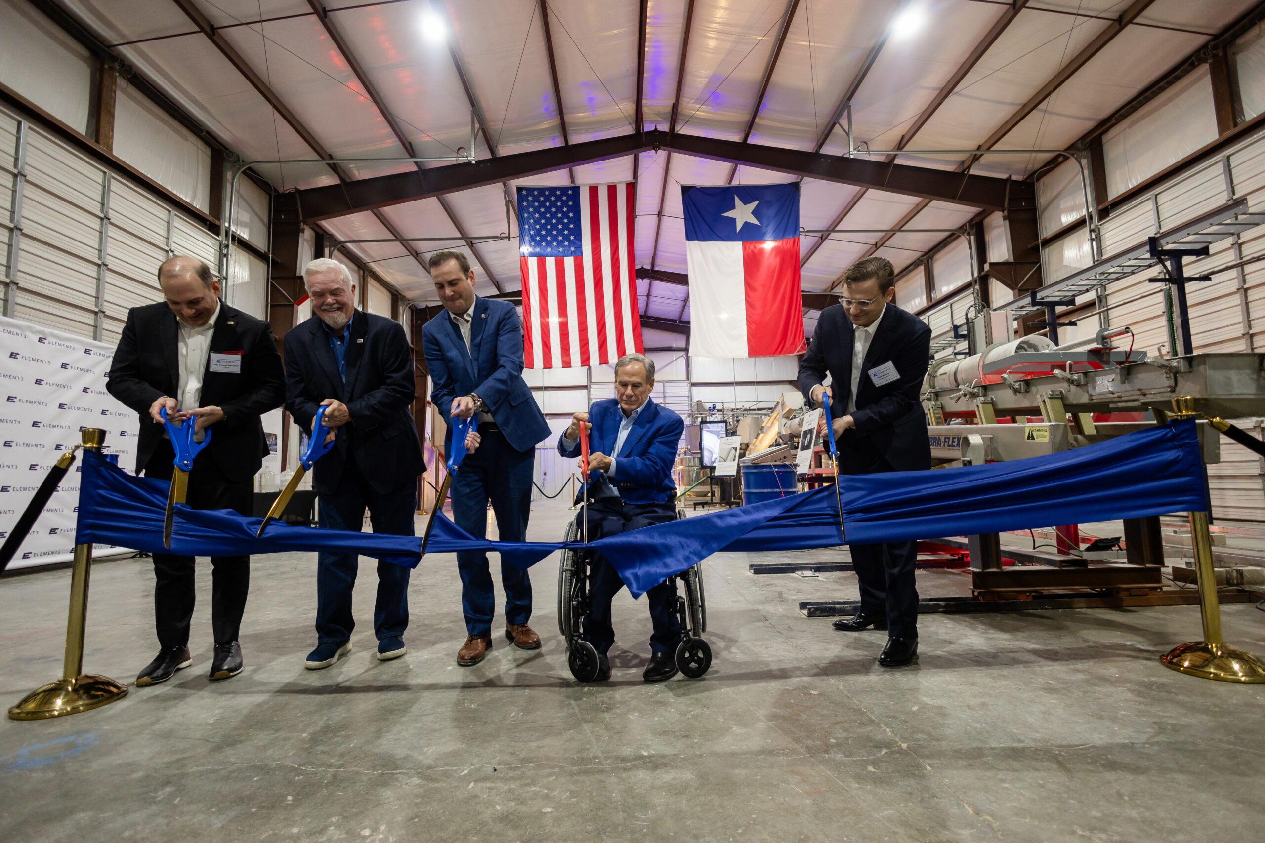 Five men in suits cut a ceremonial ribbon inside an industrial facility, with U.S. and Texas flags hanging behind them.