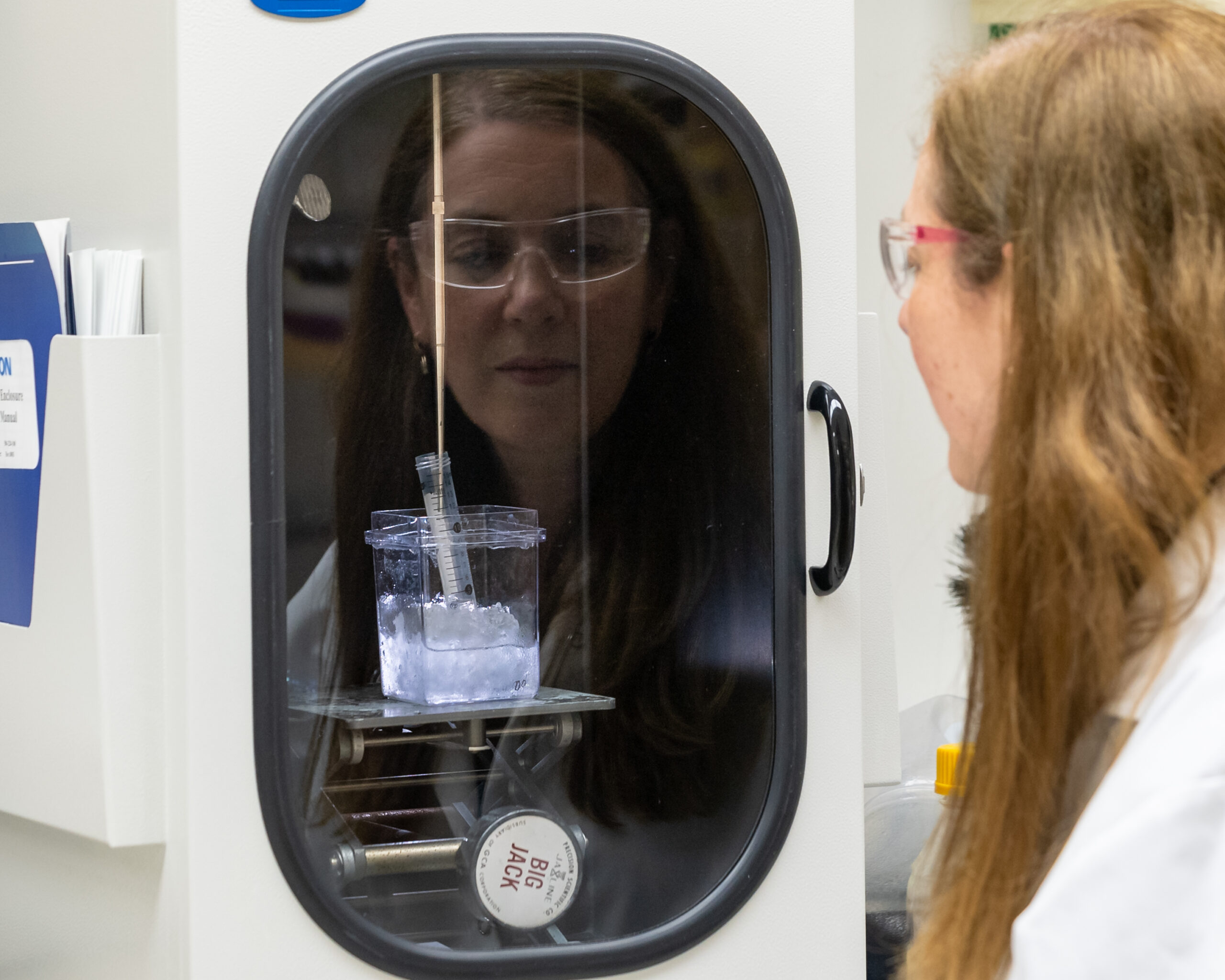 Scientist wearing safety glasses observes a syringe placed in a container of ice inside a shielded laboratory enclosure.