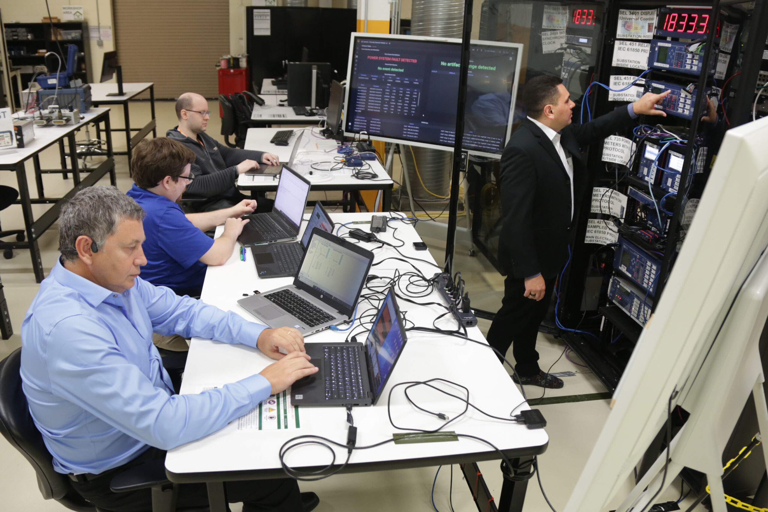 Researchers work on laptops and monitor equipment in a power systems laboratory with server racks, control displays, and testing hardware.