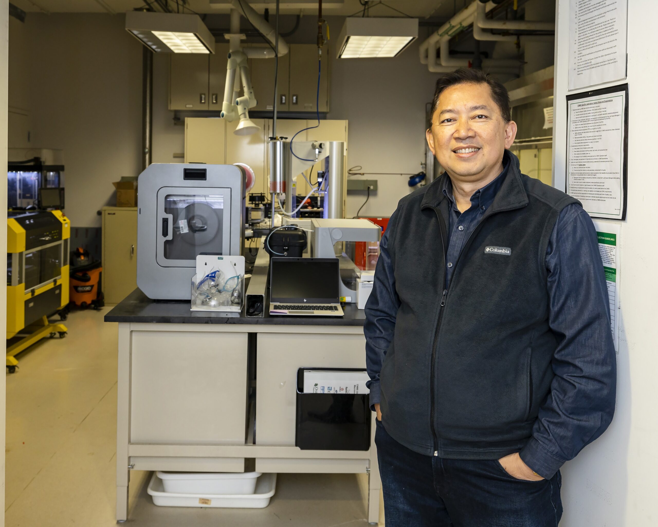 ORNL researcher is standing against the wall with the backdrop of a lab at ORNL
