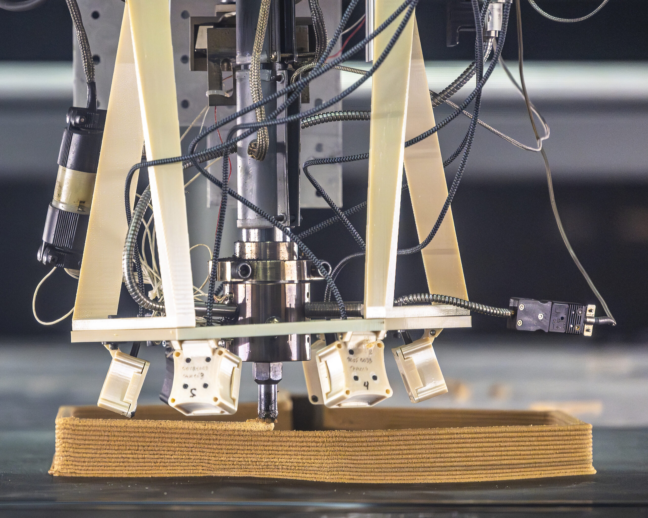 Close-up of a large-scale 3D printer depositing layers of composite plastic while surrounded by thermal cameras, sensors, and wiring used for real-time monitoring and error correction.