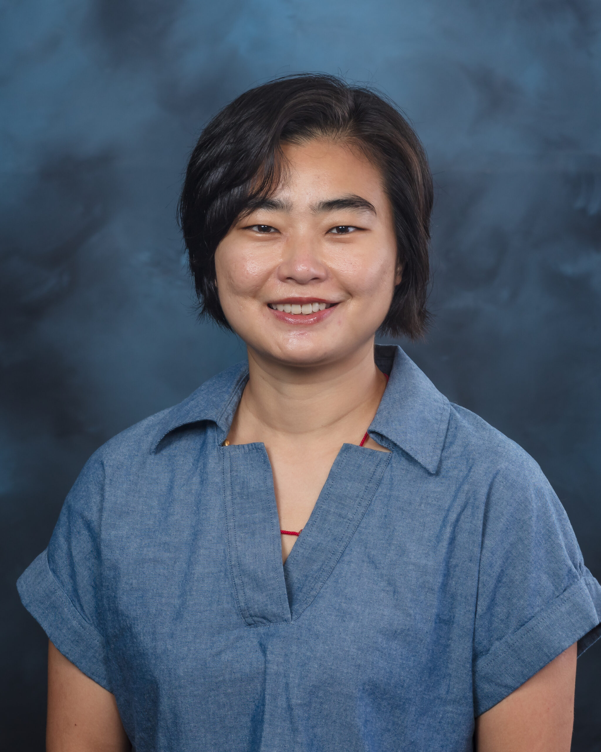 Studio portrait of a woman with short dark hair wearing a blue blouse, smiling at the camera against a blue-gray textured background.