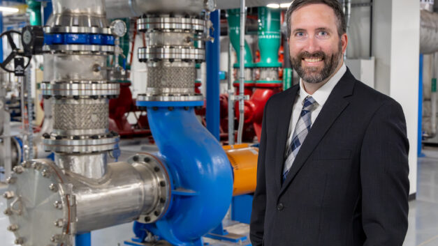 A man in a suit stands in front of large industrial piping and equipment inside a laboratory, smiling for a portrait.
