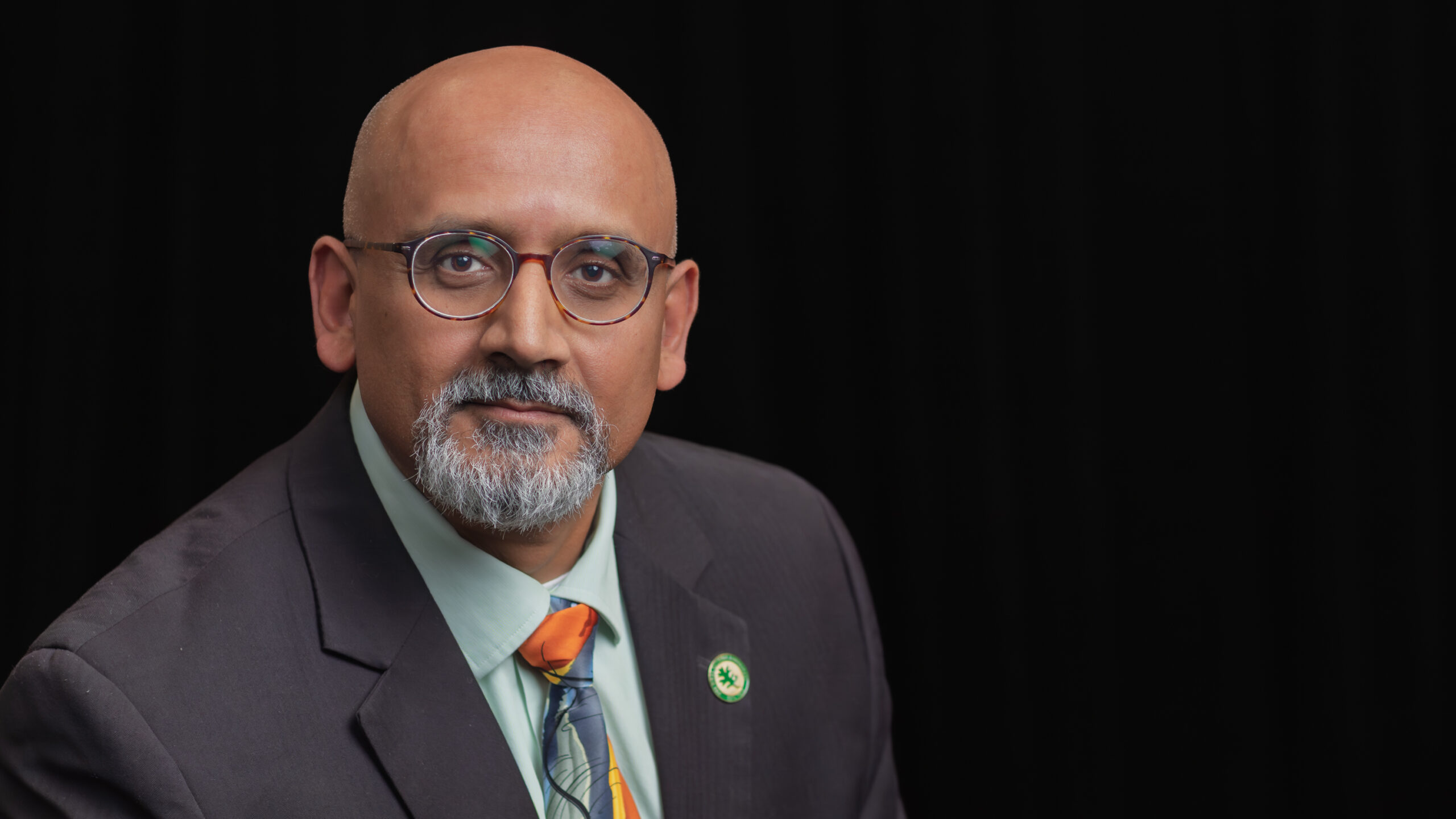 Bald man in glasses wearing a suit and colorful tie against a black background.