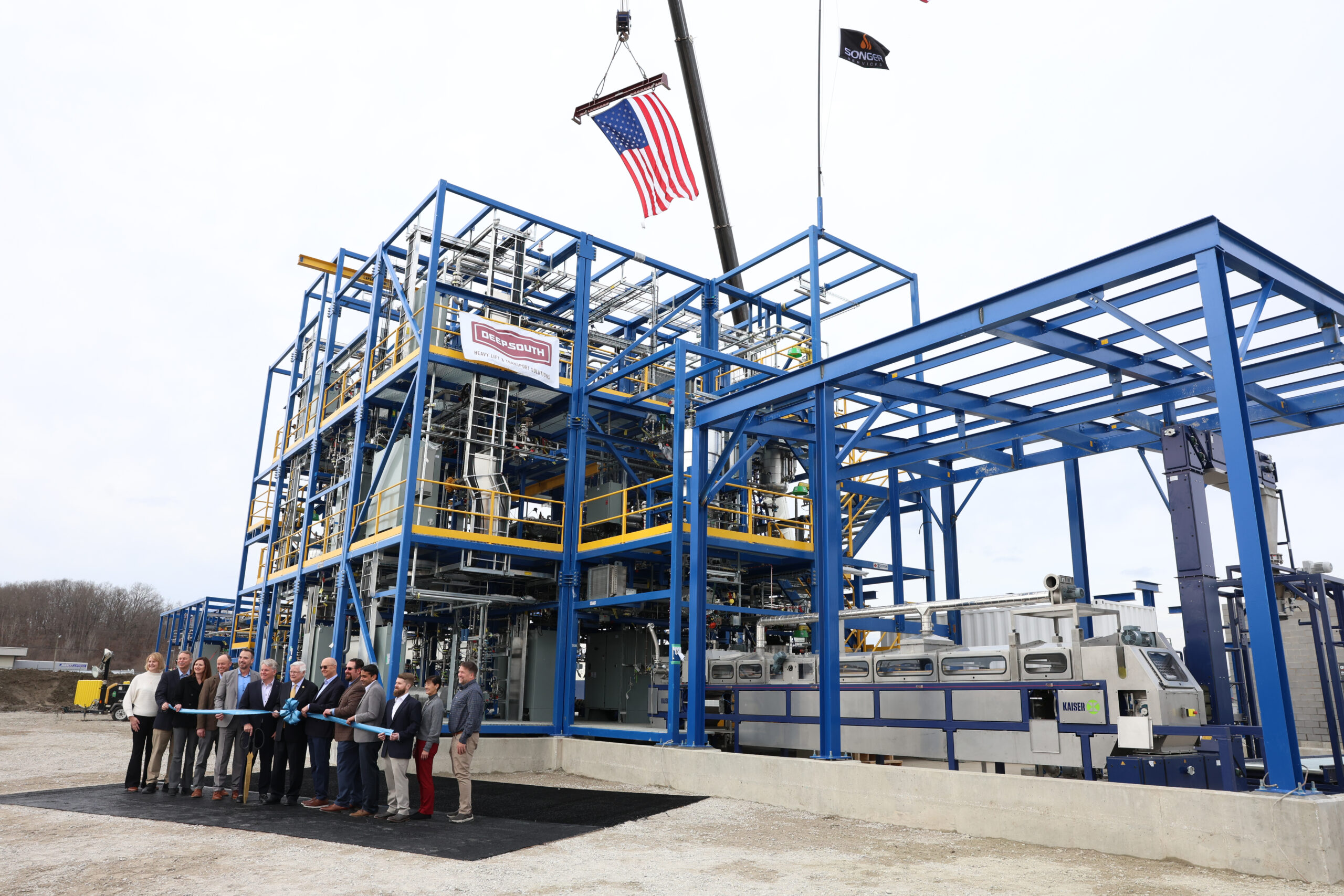 Officials gather for a ribbon-cutting ceremony in front of a newly constructed industrial pilot plant, with a large American flag suspended overhead.