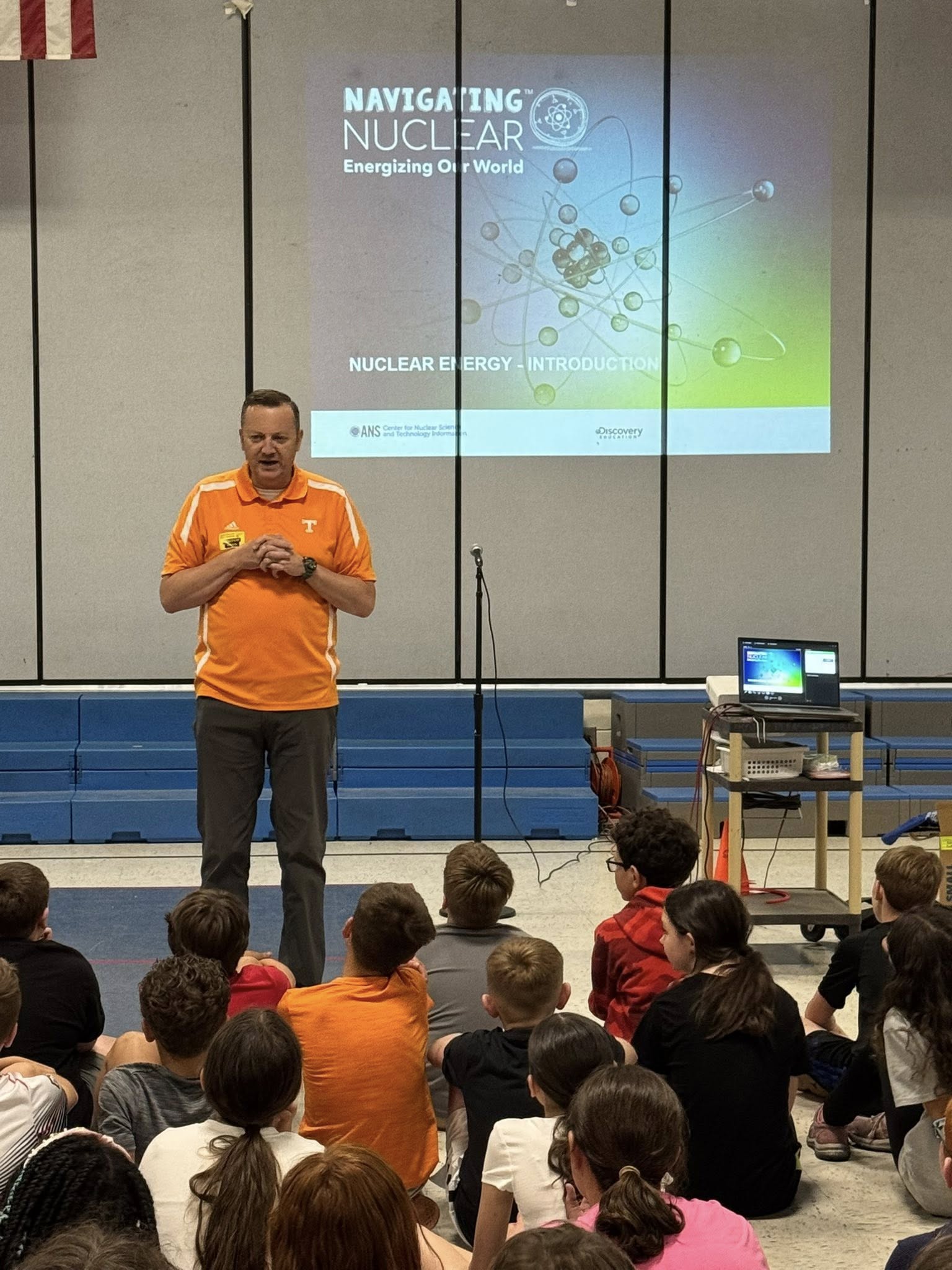 Speaker in an orange shirt presents a nuclear energy educational program to a group of students seated in a school gymnasium.