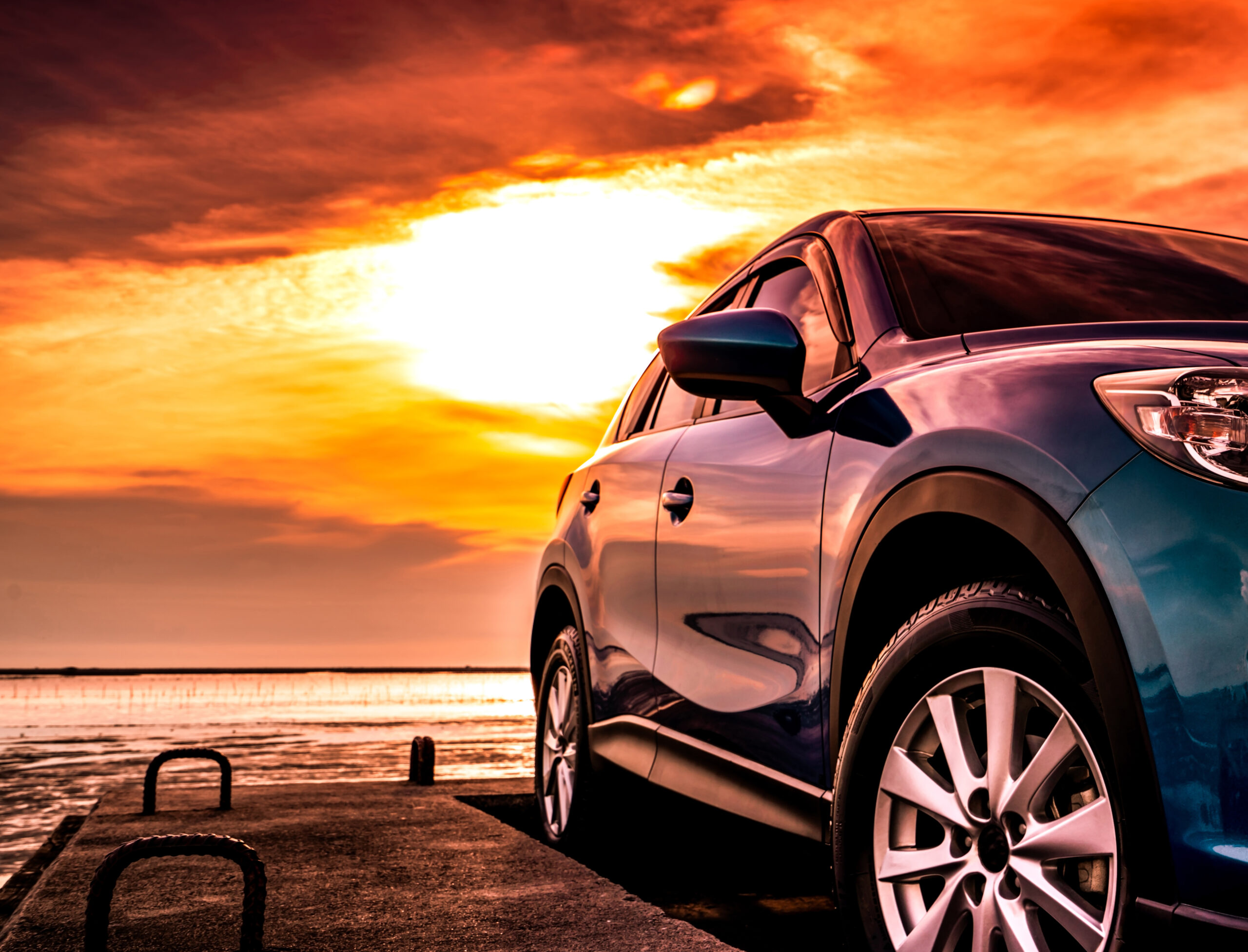 Blue SUV on a pier at sunset with vivid orange sky.