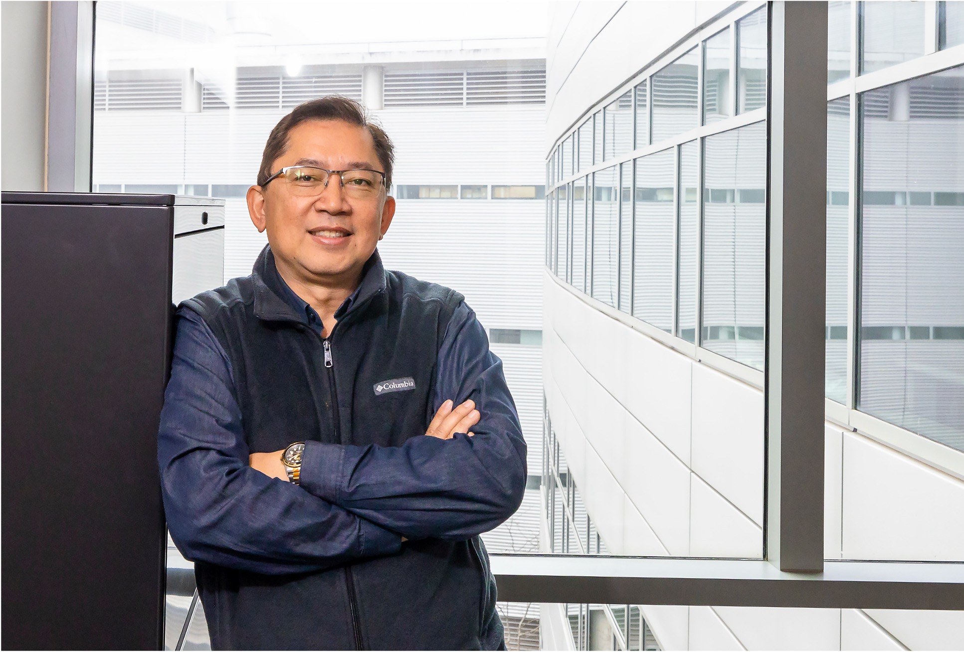 Man standing with arms crossed in a bright modern office hallway, smiling at the camera.