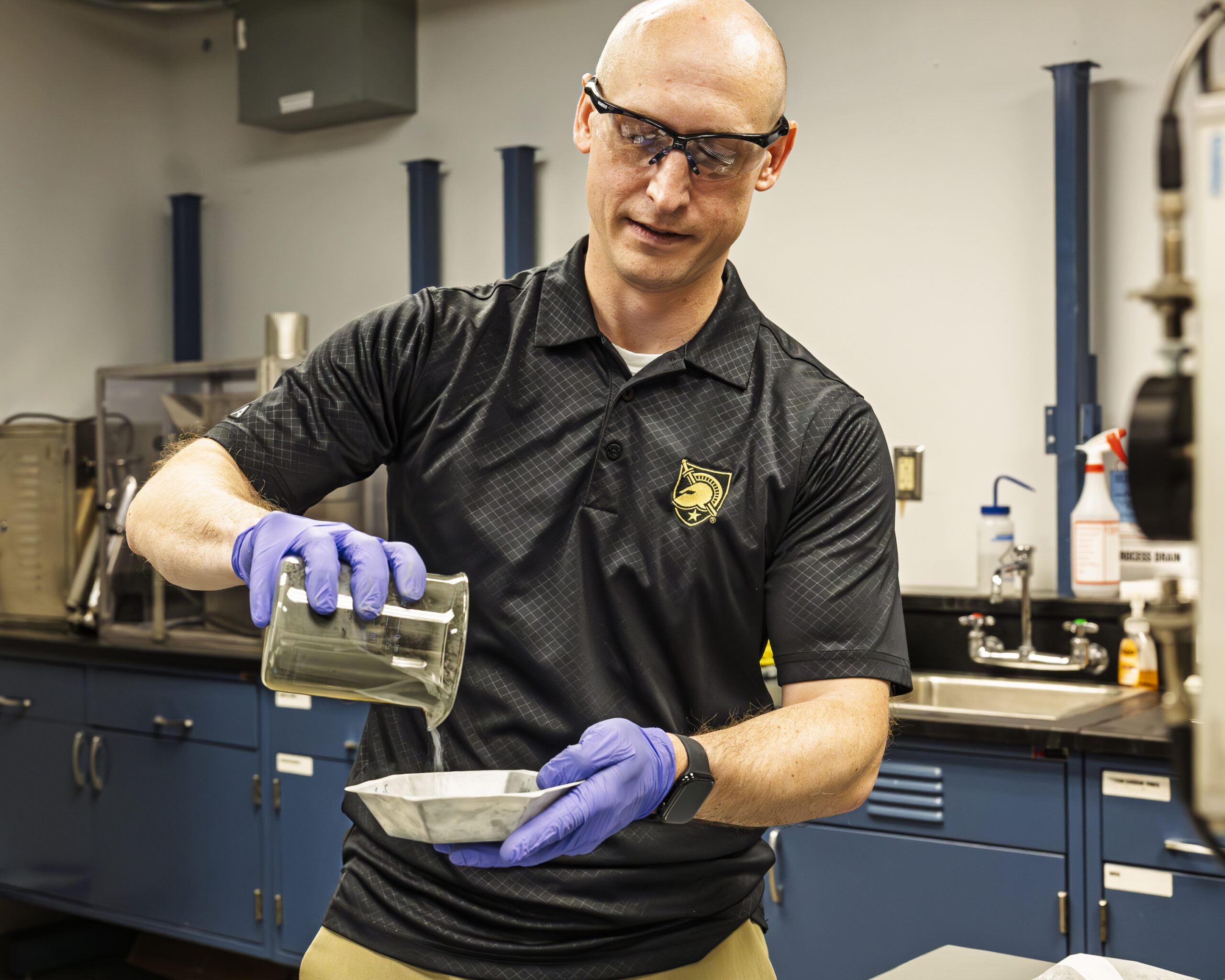 A man in safety glasses and gloves pours a liquid from a beaker into a container in a laboratory.