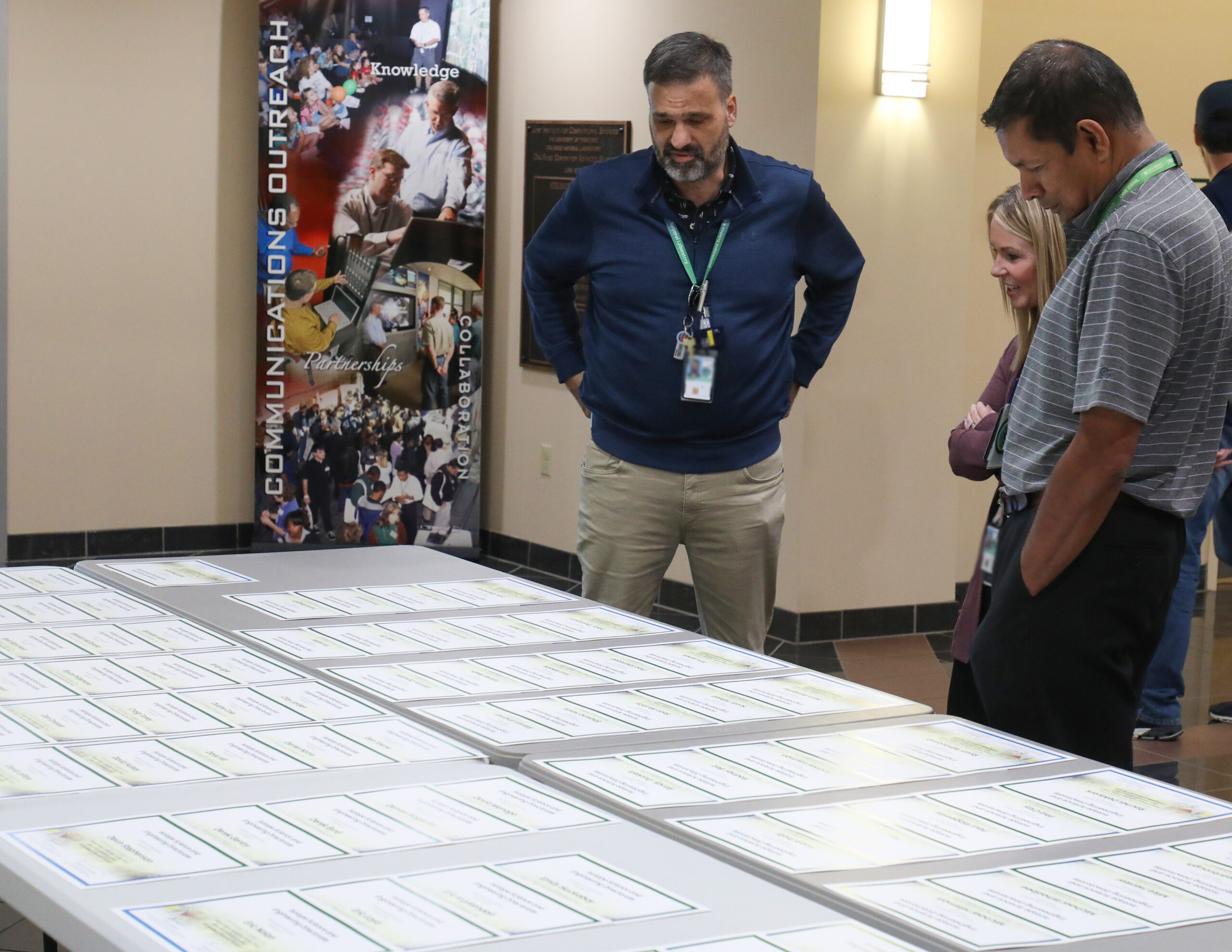 A group of professionals stand around a large table reviewing printed materials laid out in rows, engaged in discussion inside a building lobby or conference area.