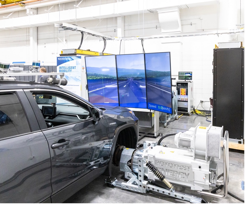 A gray SUV is mounted on a chassis dynamometer inside a laboratory, with its front wheels secured to testing equipment labeled “Rototest Energy.” In front of the vehicle, three large screens display a simulated roadway scene with a speed readout of 0.0 km/h, indicating a driving simulation or vehicle testing setup. Cables, control cabinets, and lab equipment surround the test area in a clean, industrial research facility.