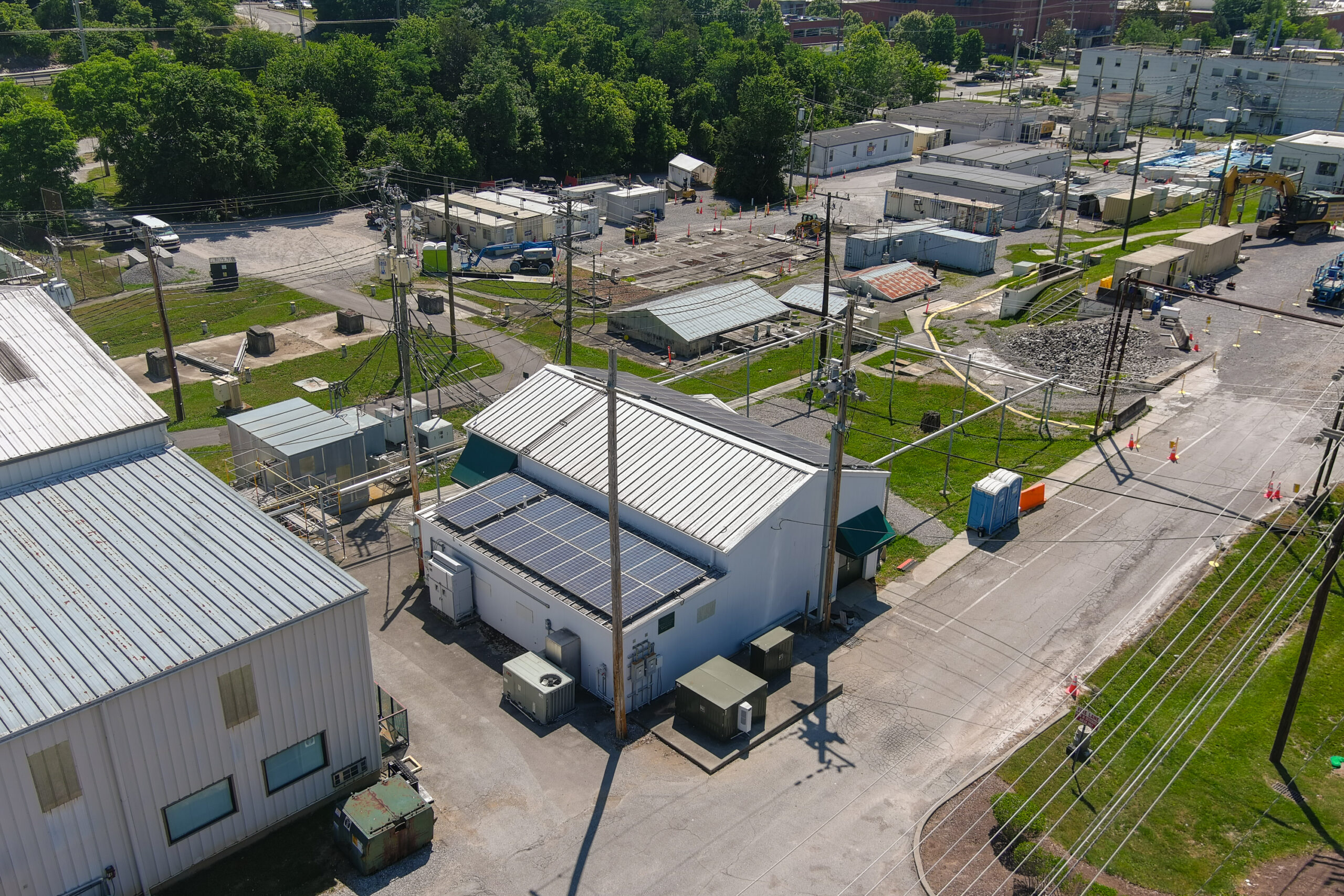 Aerial view of an industrial utility site with multiple small buildings, electrical equipment, and utility poles. A white building in the foreground has rooftop solar panels, surrounded by transformers, cables, and service boxes. The area includes paved access roads, gravel lots, construction equipment, and temporary containers, with trees and additional industrial buildings in the background.