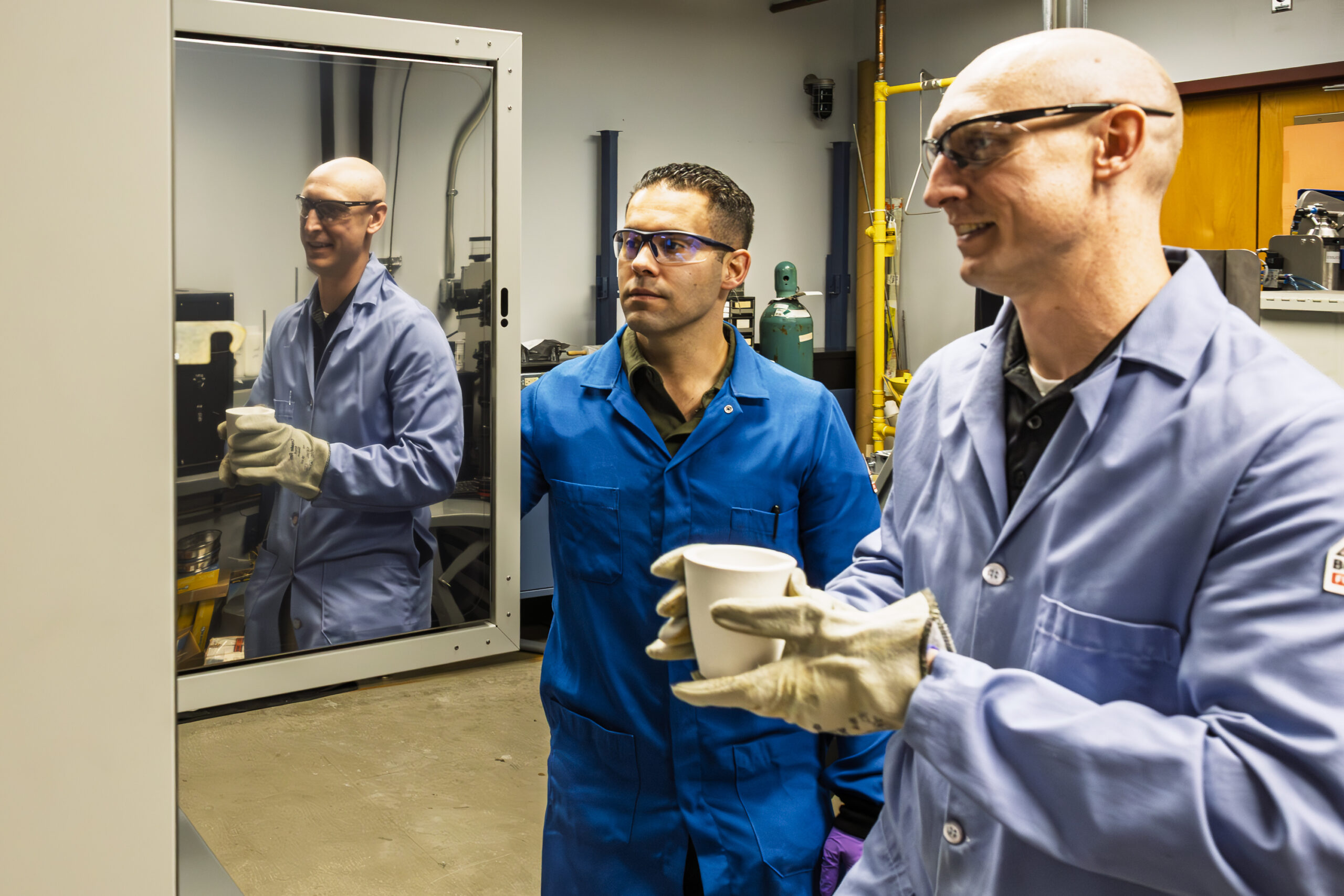 Two men wearing lab coats and protective eyewear observe a sample inside a lab, standing near a glass enclosure.