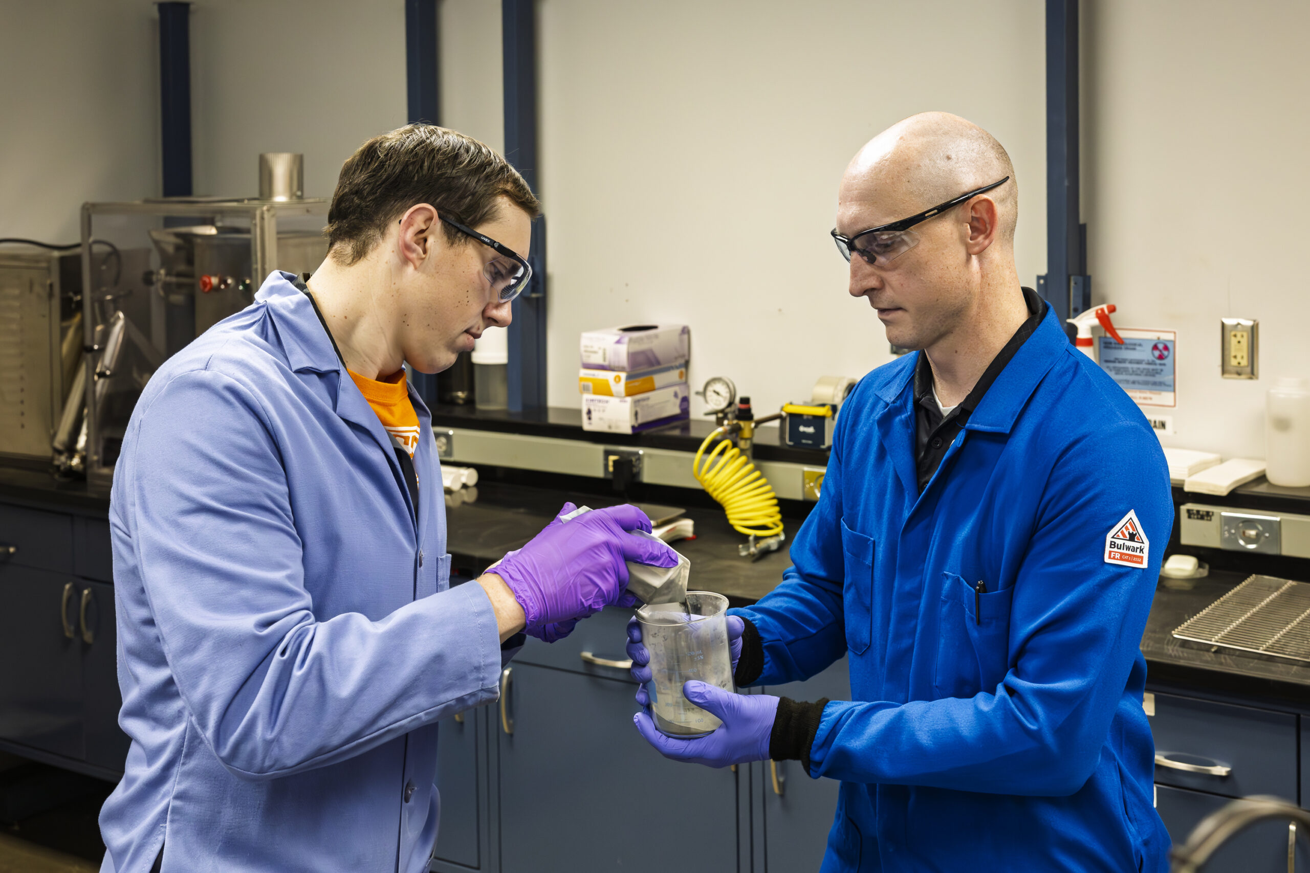 Two men in lab coats and safety glasses examine and pour a sample into a beaker in a lab setting.