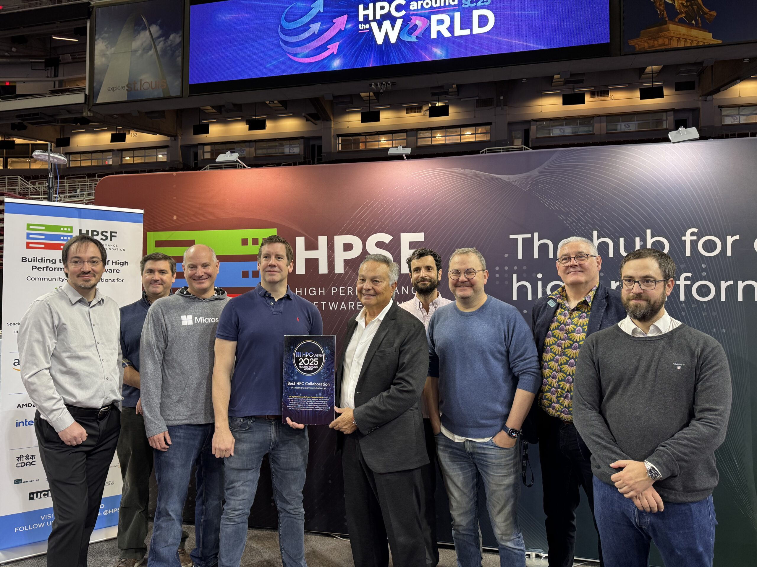 A group of ten people stand together at an HPC conference booth, posing for a photo while holding a glass award plaque. Behind them are banners reading ‘HPSF’ and ‘The hub for high-performance software,’ and a large overhead sign that says ‘HPC around the World.’ The group appears to be celebrating an HPCwire 2025 Best HPC Collaboration award.